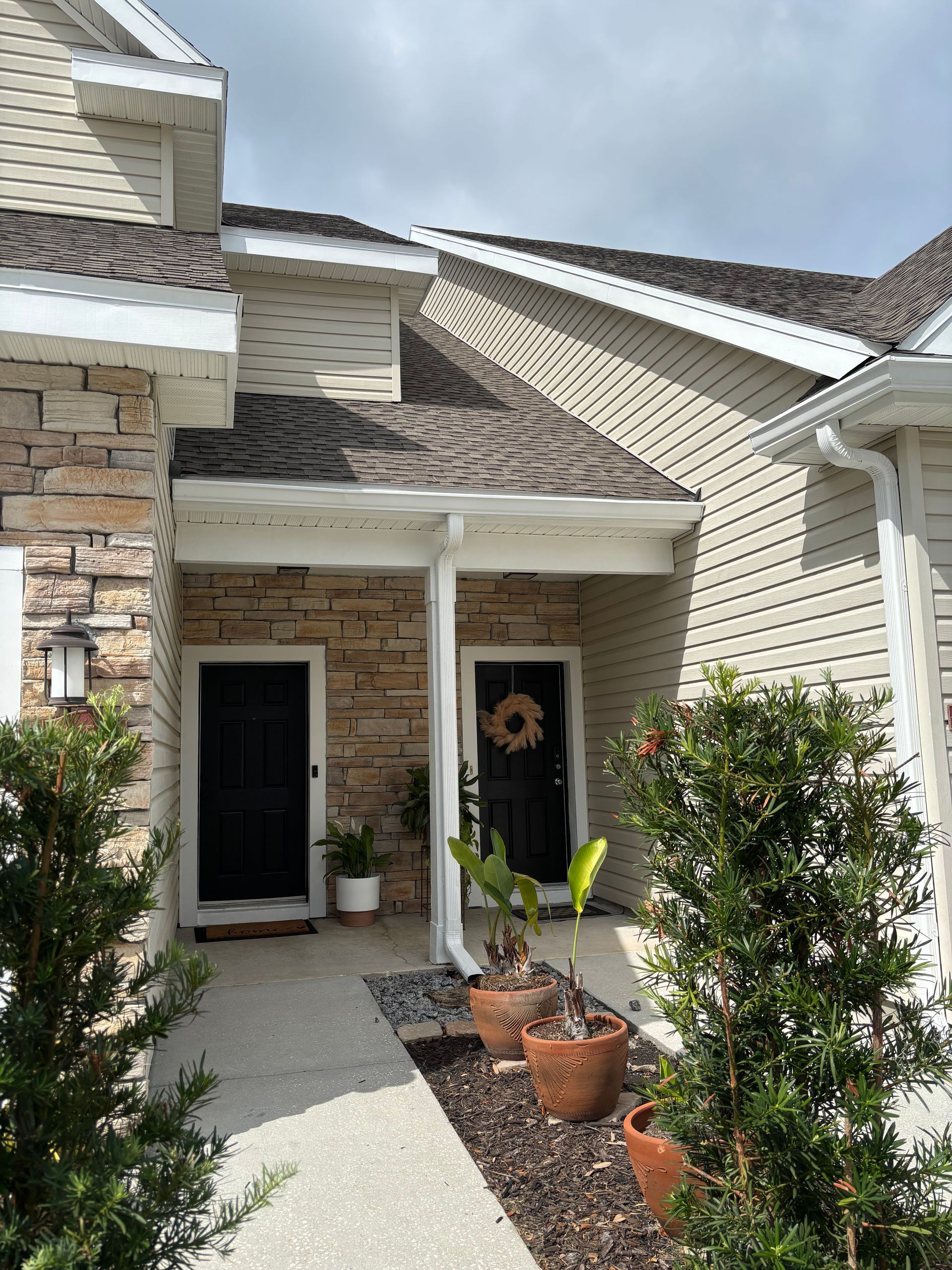 Front porch with stone columns, white siding, potted plants, and a dark front door under a gabled roof.