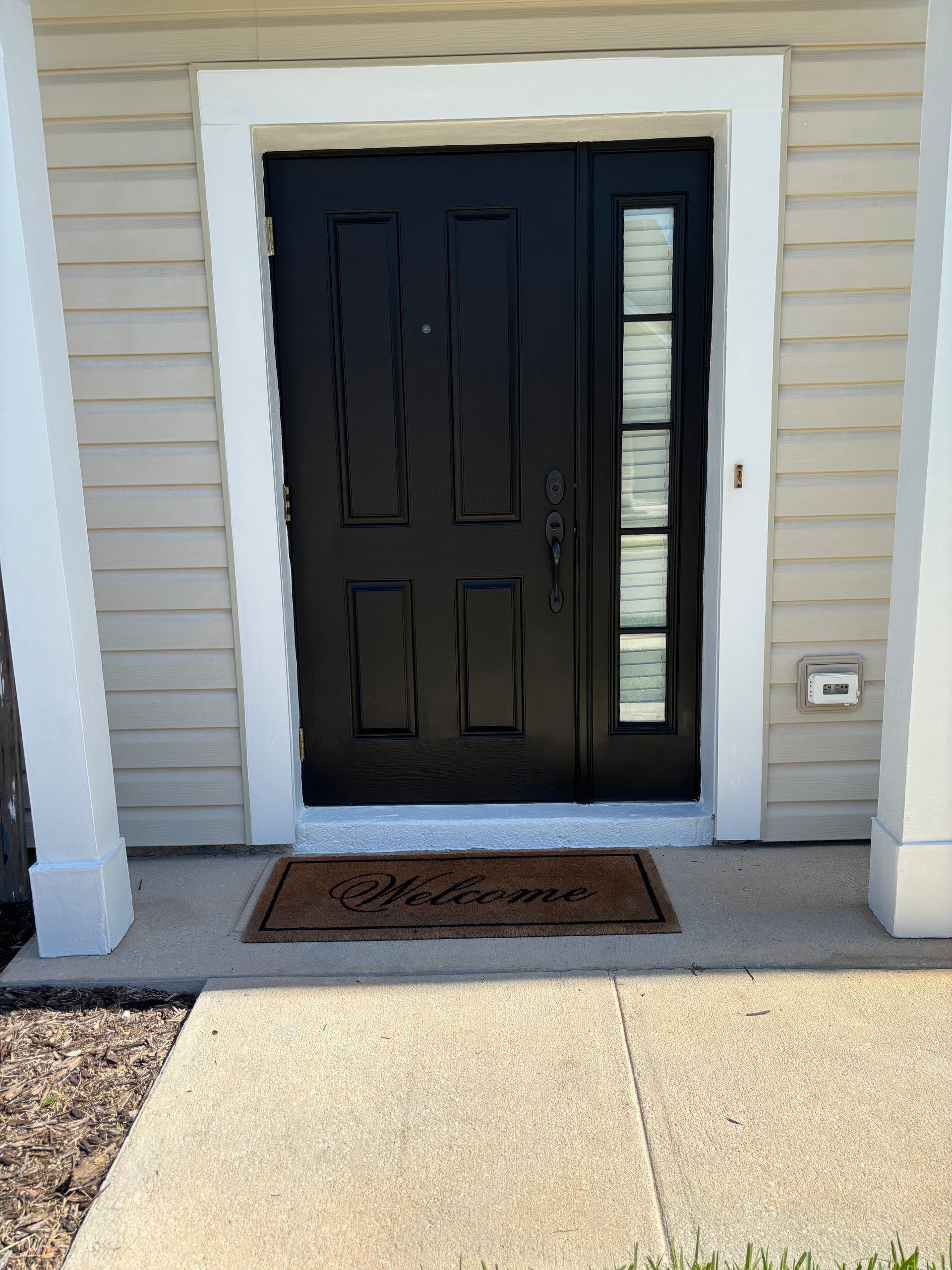 Front entry with black door, narrow side windows, and a welcome mat on a concrete porch.