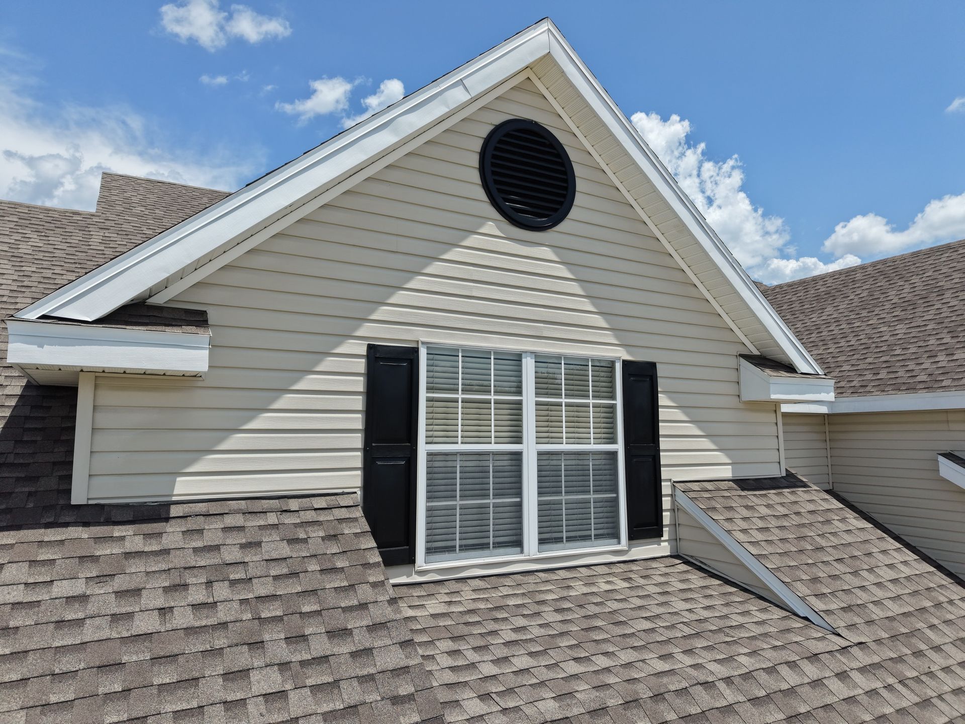 Suburban house roof with beige siding, black shutters, and a round attic vent against a blue sky