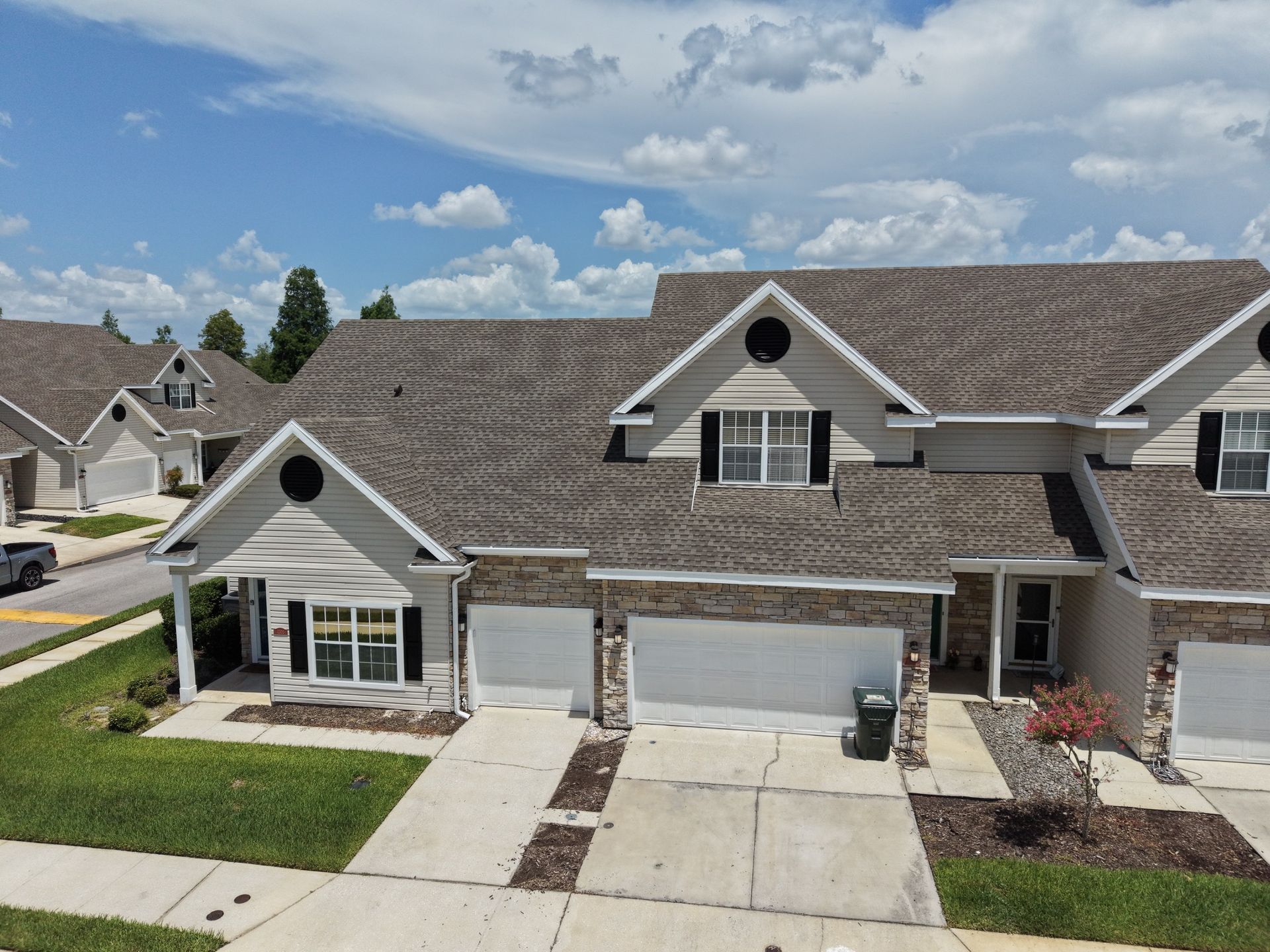 Suburban townhouse with gray roof, white garage doors, front lawn, and paved driveway under a partly cloudy sky