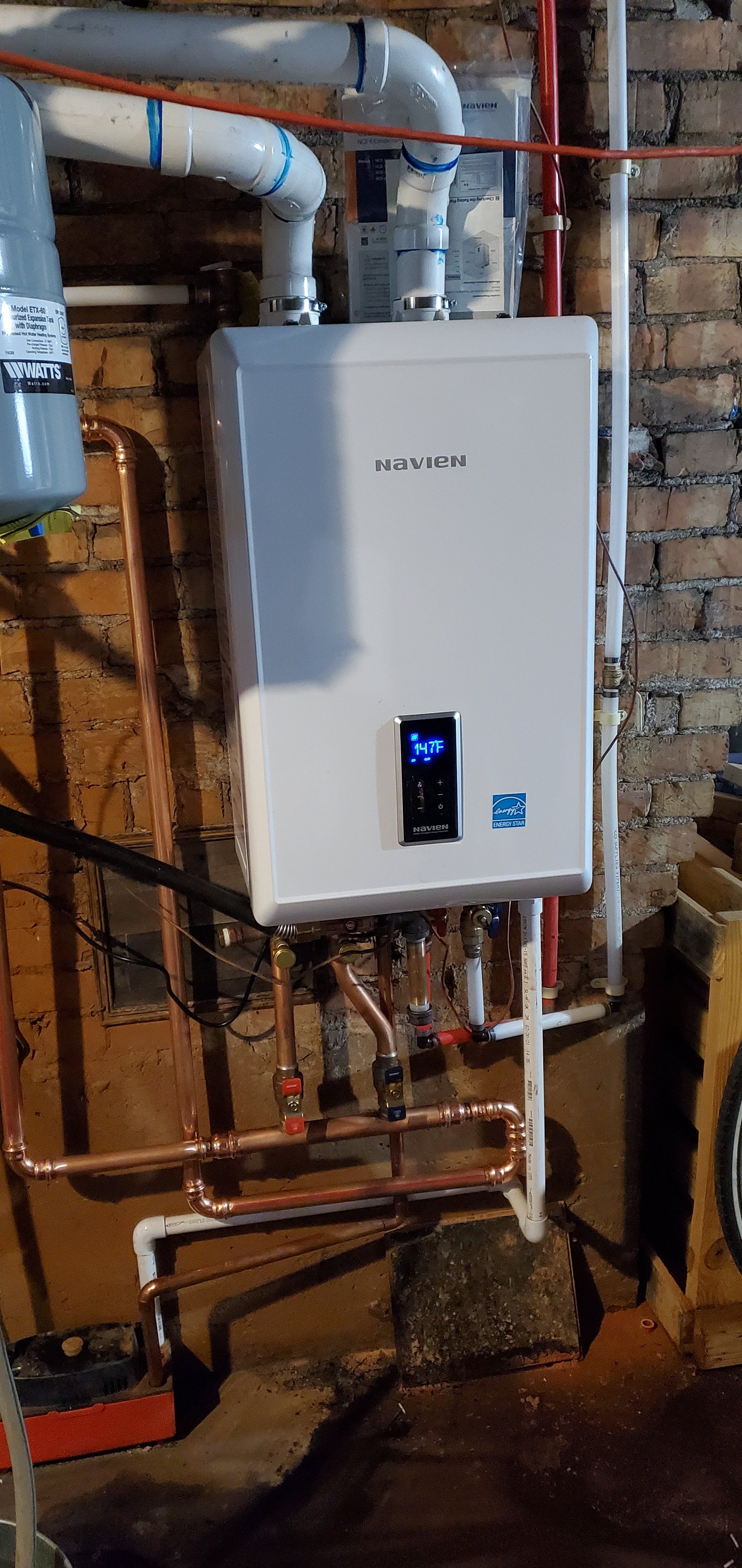 A white water heater is mounted to a brick wall in a basement.