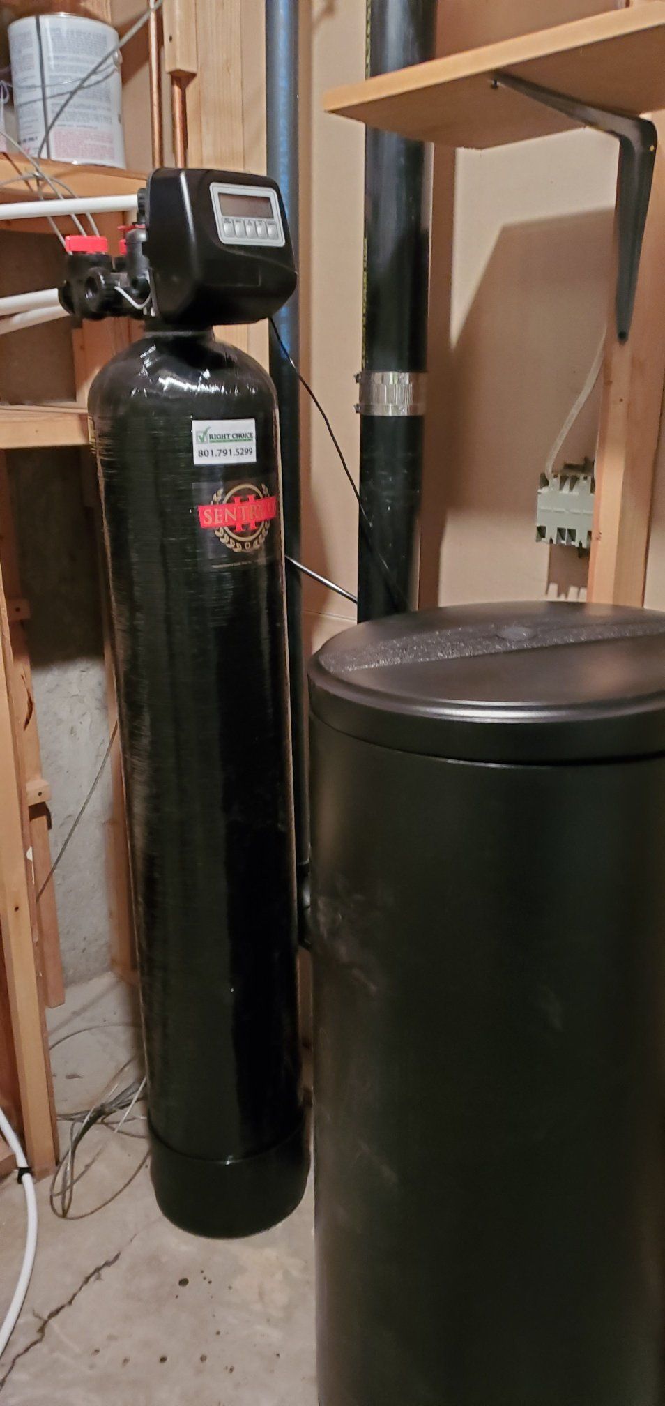 A water softener is sitting in a basement next to a trash can.