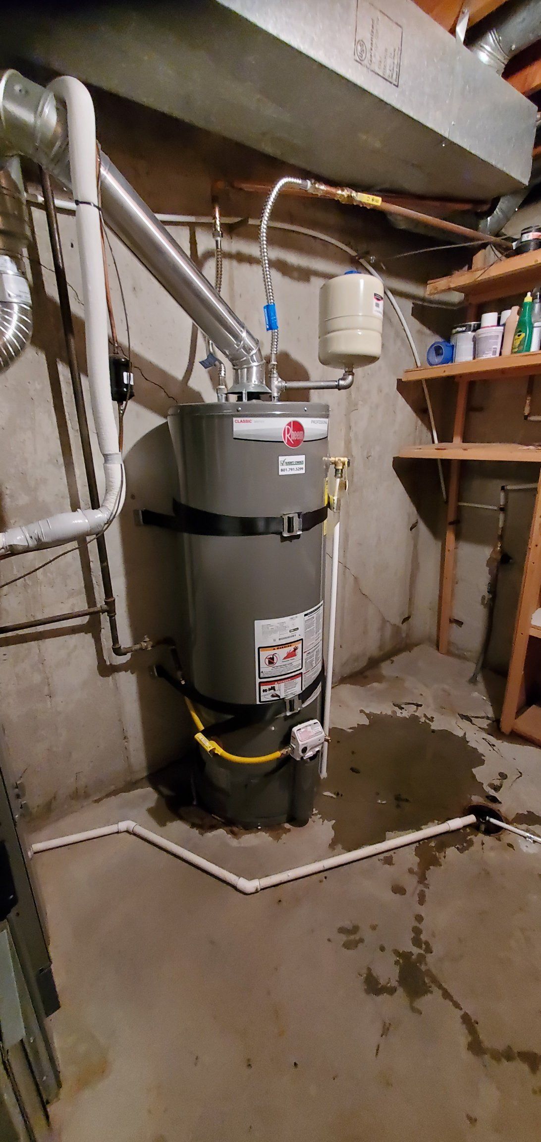 A water heater is sitting in a basement next to a wooden shelf.