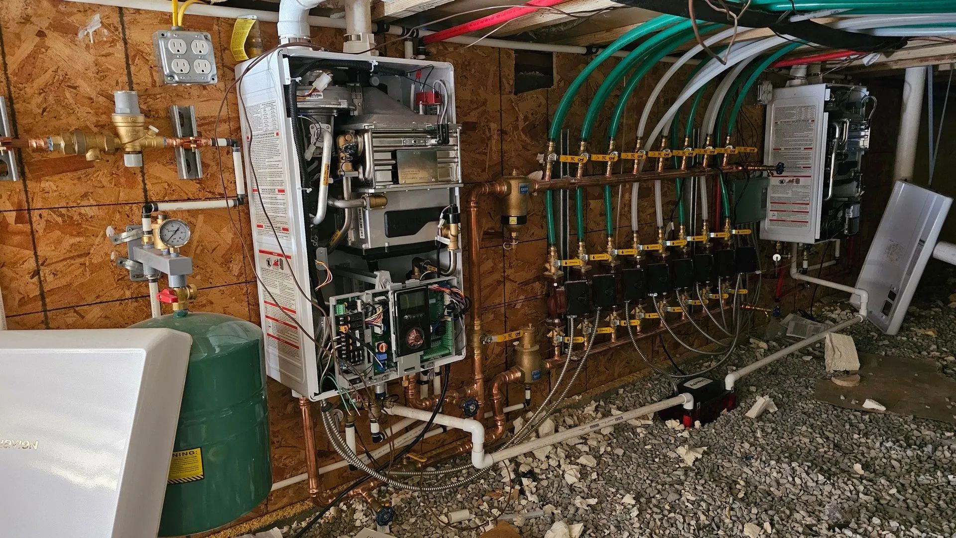 An exposed tankless water heater connected to a manifold of pipes and electrical equipment mounted on a wooden wall.