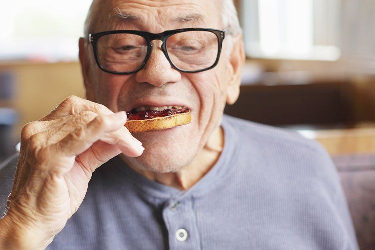 Elderly person wearing glasses eating a cookie in a bright indoor setting