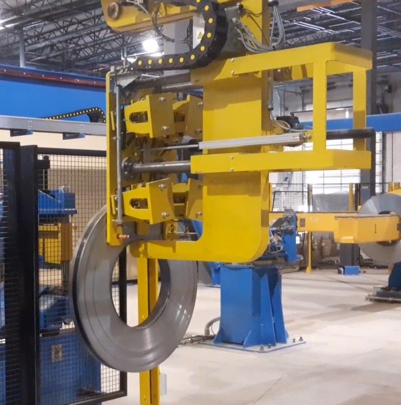 Braccio robotico giallo che solleva una grande bobina di metallo in un ambiente industriale.
