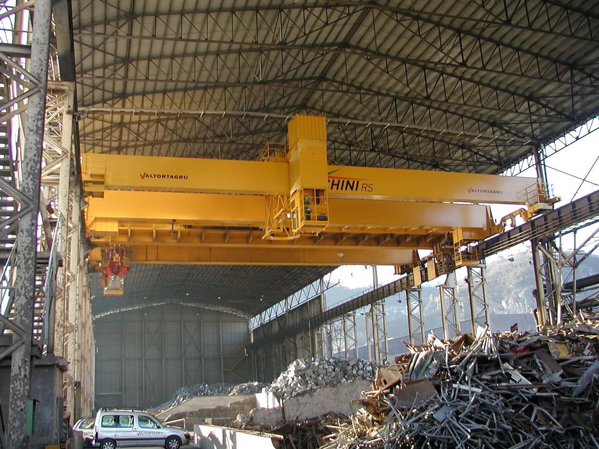 Gru a ponte gialla che solleva rottami metallici in un magazzino.