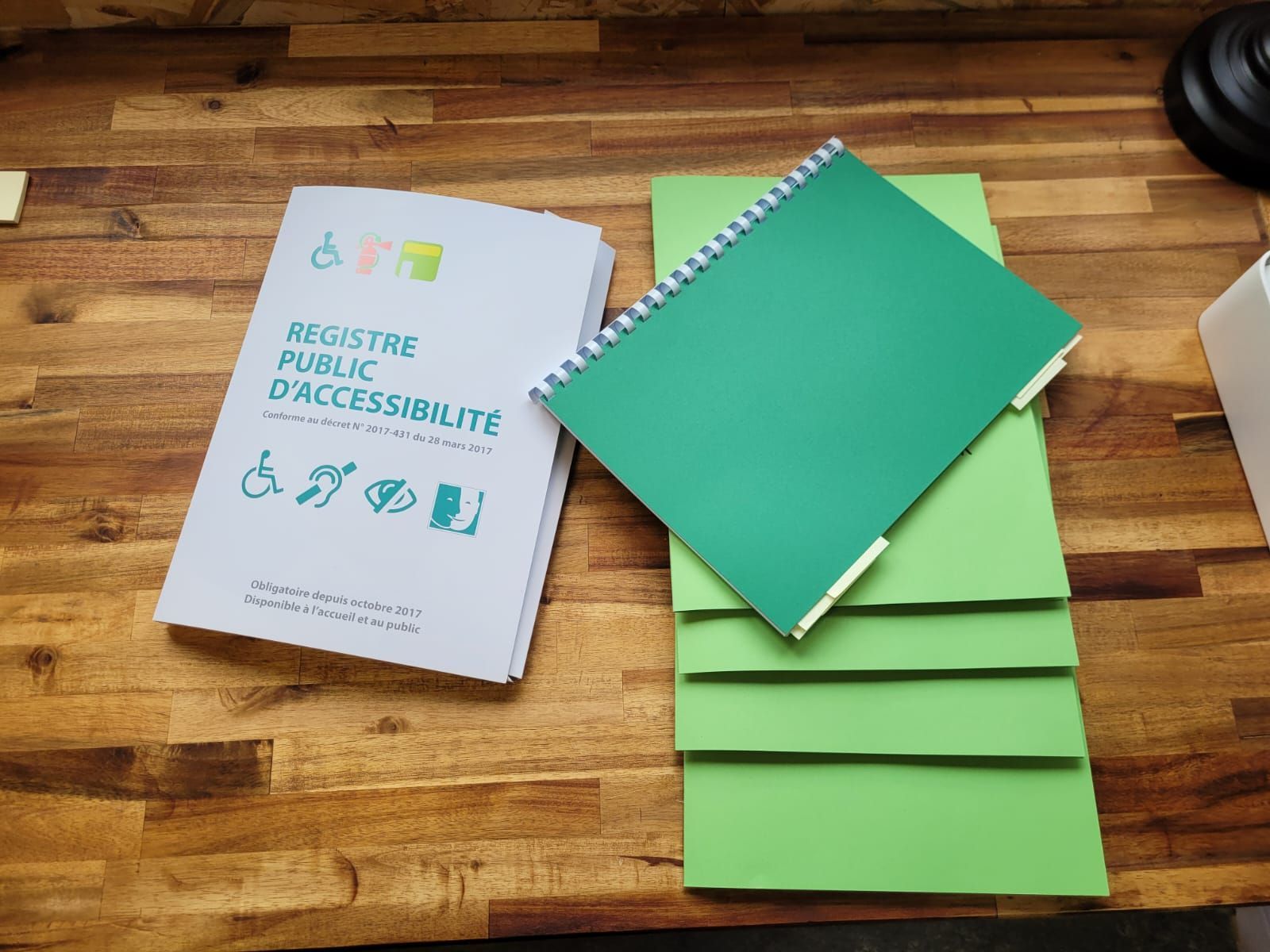 Pile de livrets blancs et un cahier à spirale vert sur une surface en bois, étiqueté «Registre public d'accessibilité».