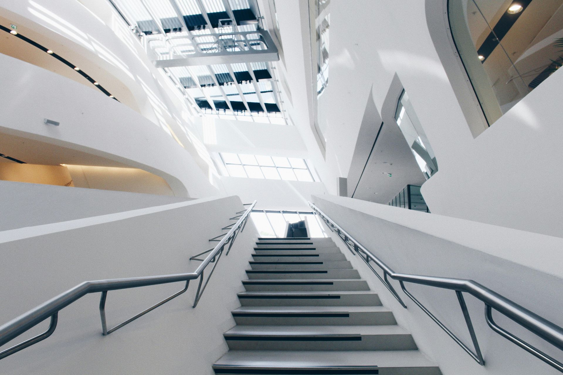 Intérieur d'un bâtiment moderne avec un large escalier menant à l'étage. Murs blancs, rampes en acier et plafond en verre.