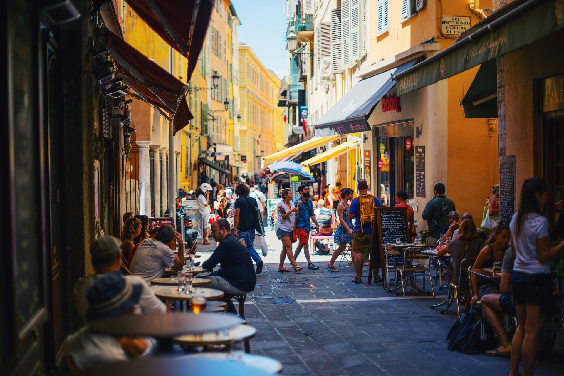 Scène de rue européenne animée, avec des gens qui marchent et déjeunent aux terrasses des cafés.