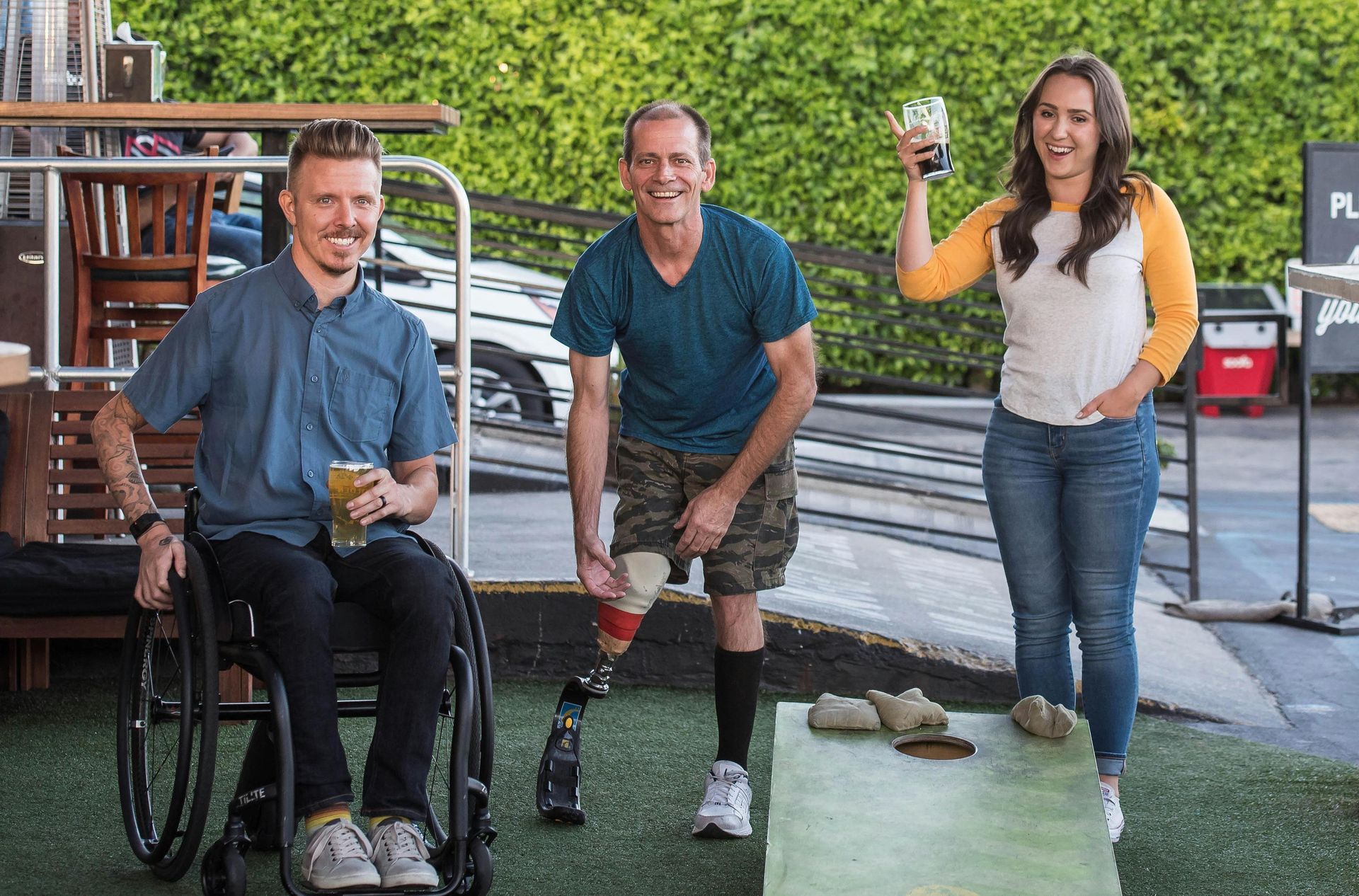 Trois personnes, l'une en fauteuil roulant et l'autre avec une prothèse de jambe, lèvent leurs verres de bière à l'extérieur.