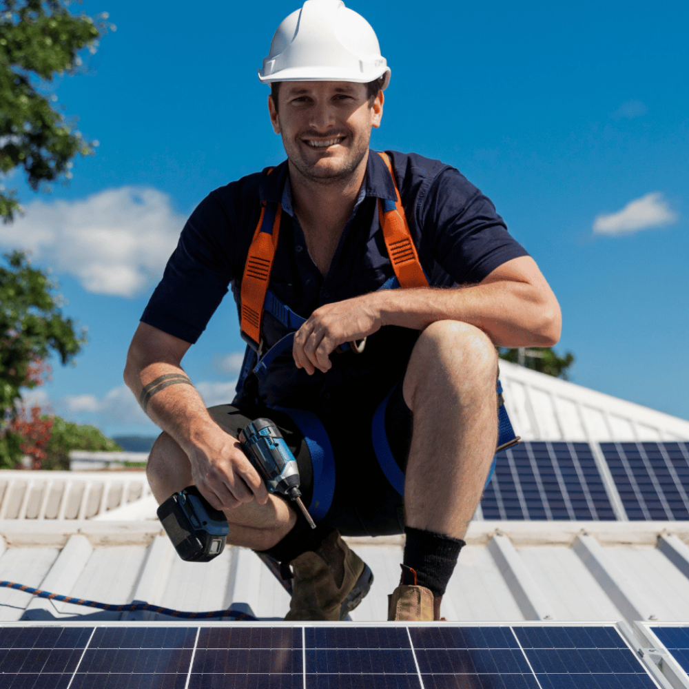 Worker kneeling on solar panels holding a power tool under a blue sky