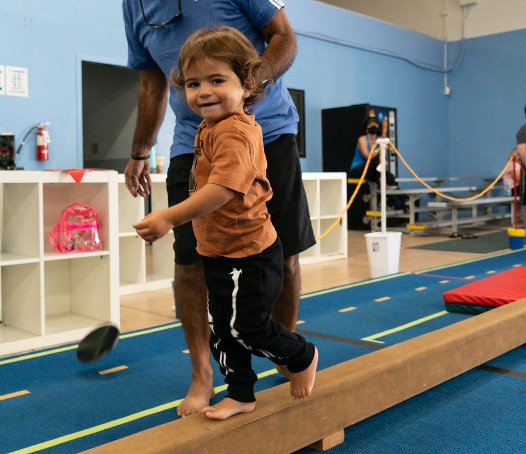 A little boy is walking on a balance beam with a man standing behind him