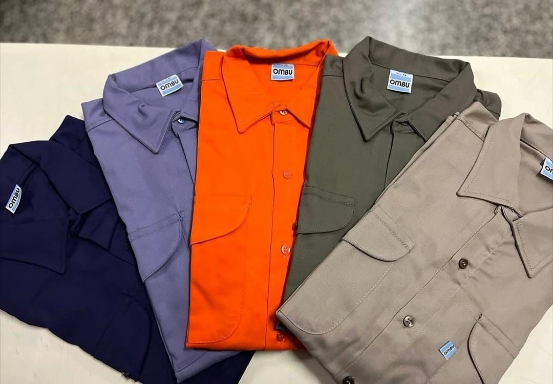 Cinco camisas de trabajo dobladas en azul marino, lavanda, naranja, verde oliva y beige, expuestas una al lado de la otra.