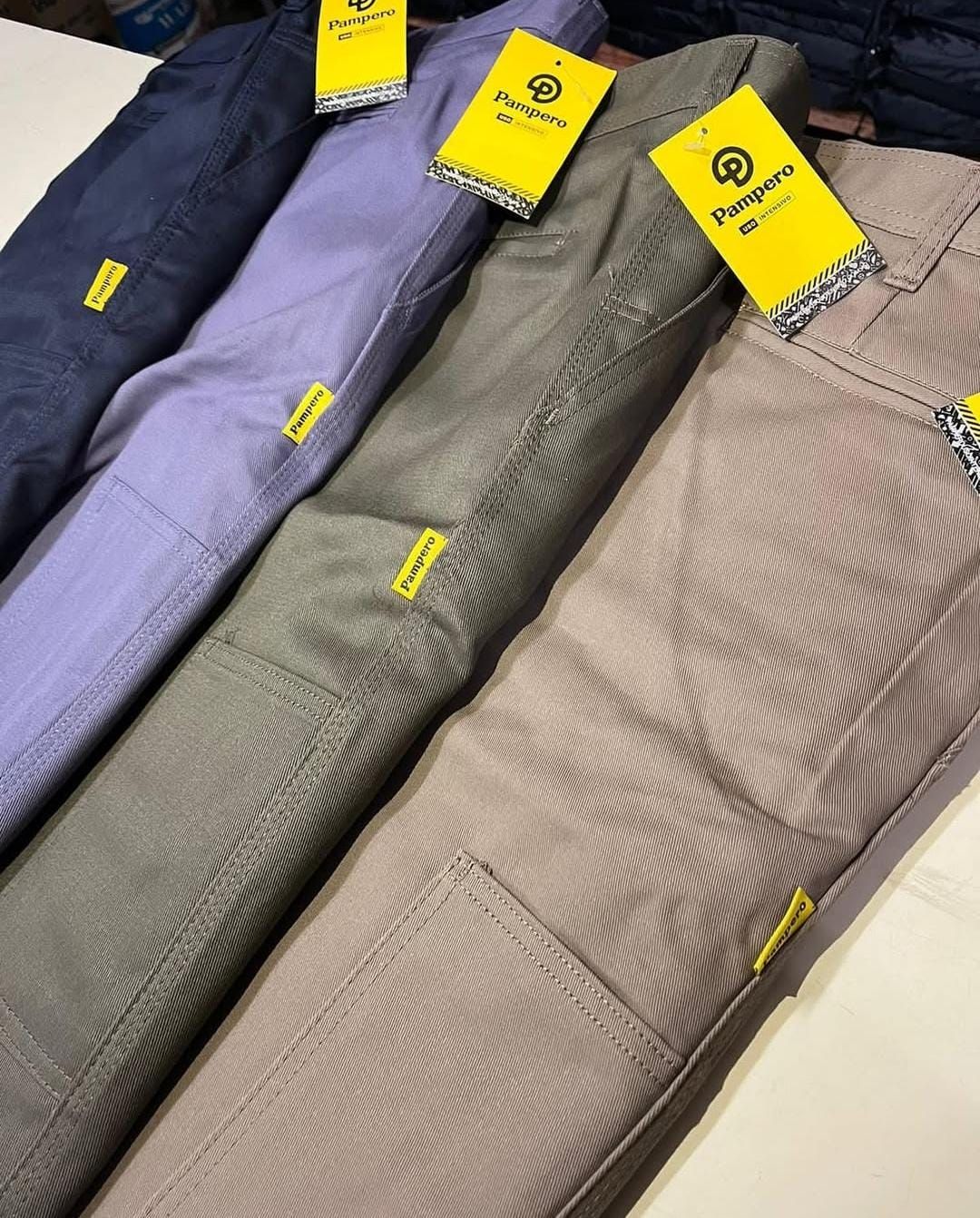 Pantalones doblados de color azul marino, lavanda, verde oliva y beige con etiquetas amarillas de la marca, expuestos sobre una mesa.