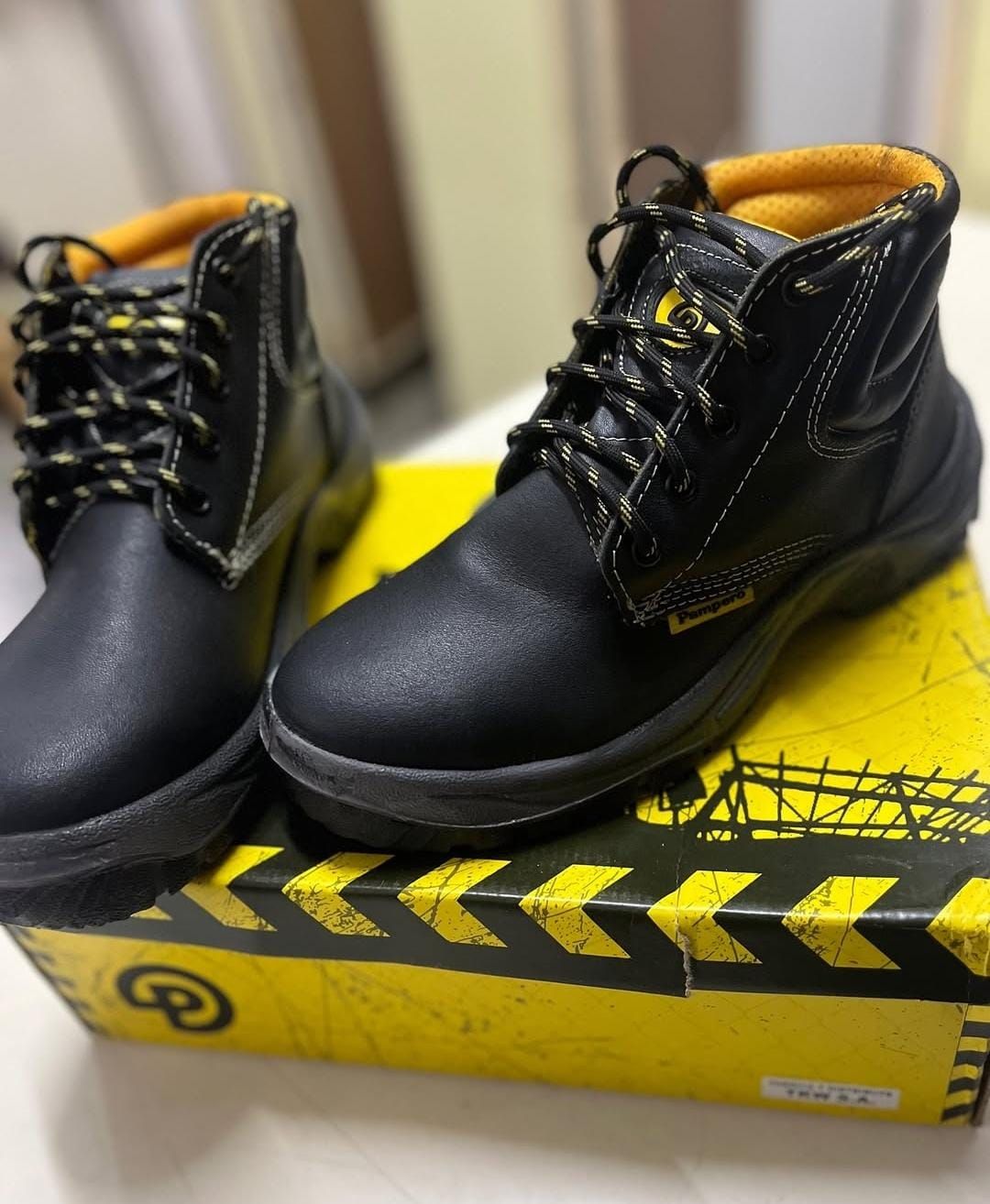 Botas de trabajo negras con cordones amarillos sobre una caja de zapatos amarilla con franjas negras de precaución.