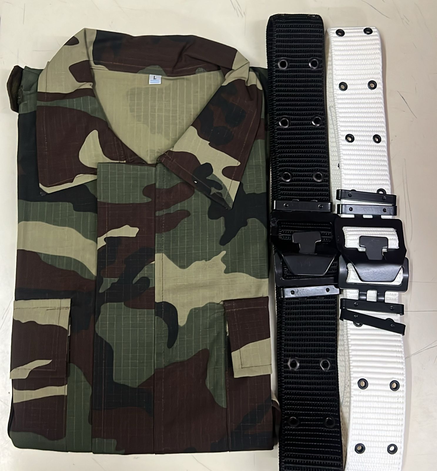 Camisa de camuflaje junto a cinturones o equipo táctico de color negro, blanco y marrón claro sobre una superficie clara.