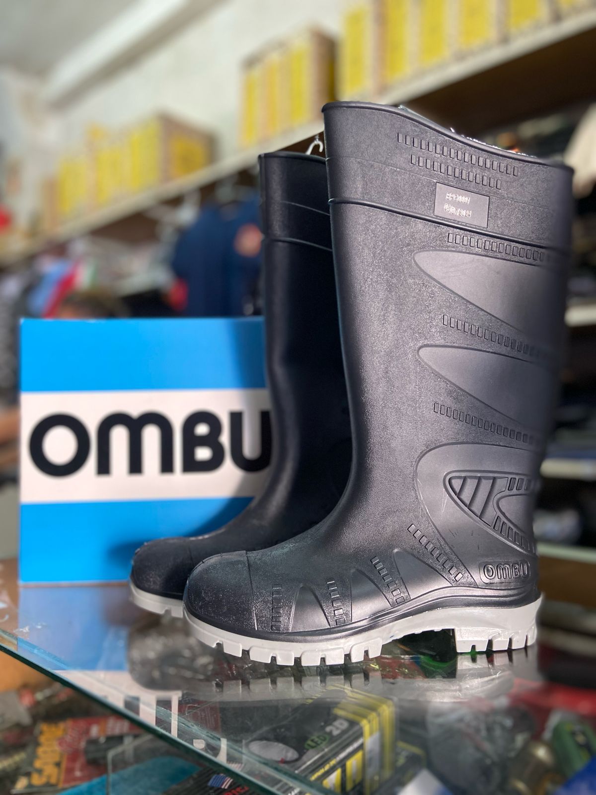 Botas de goma negras de la marca OMBU en un estante de una tienda, con el embalaje azul de OMBU detrás.