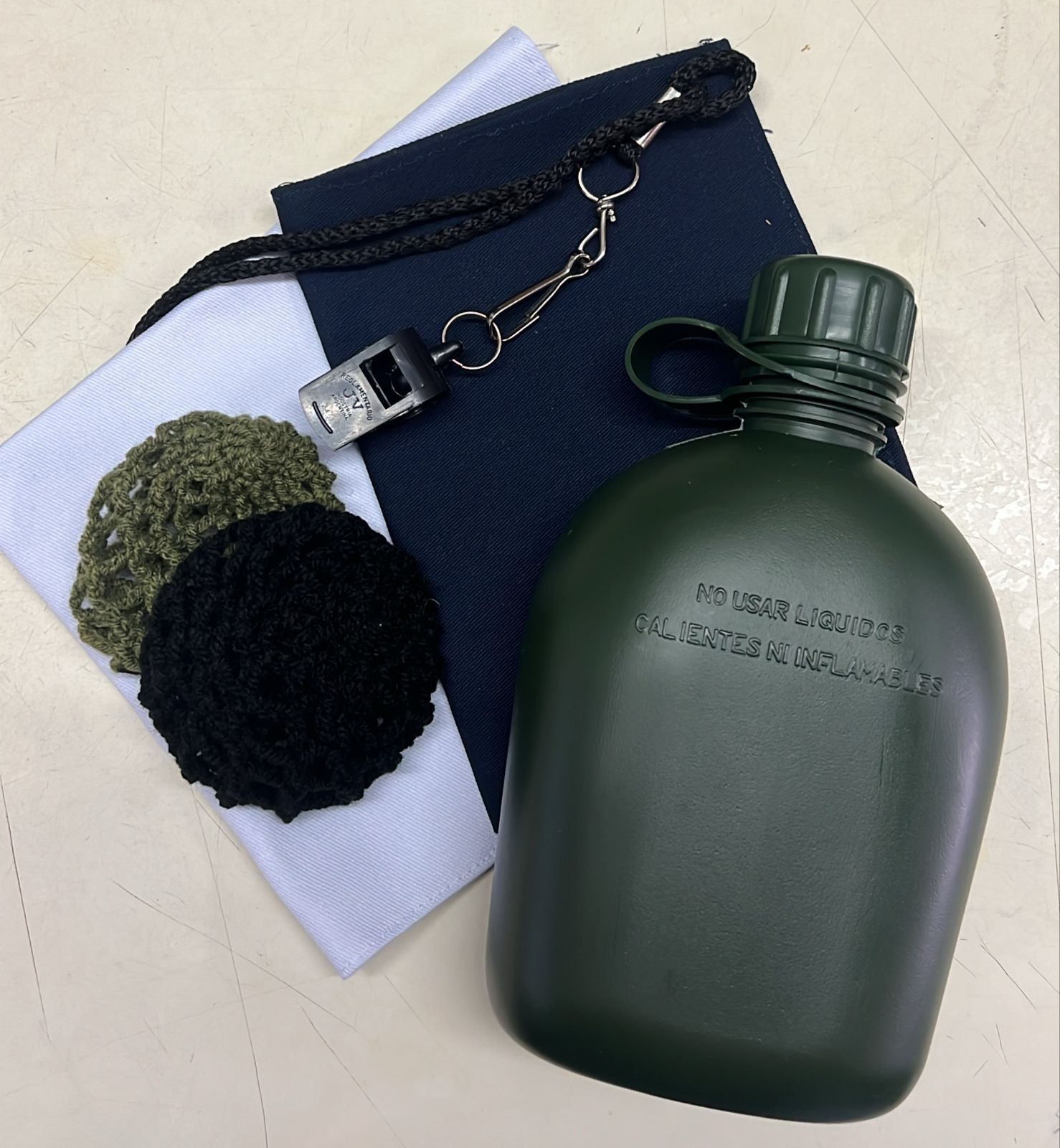 Cantimplora militar, paños doblados y un cordón con un pompón sobre una superficie blanca.