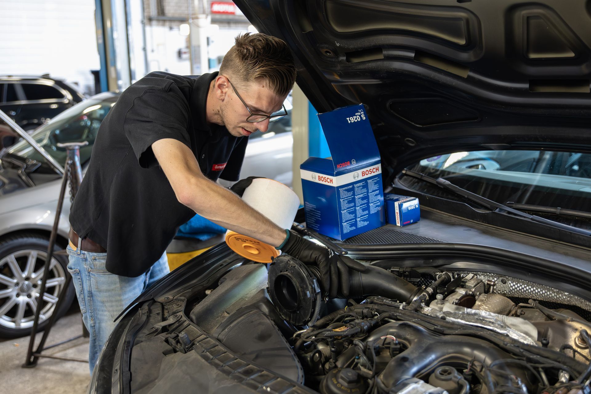 Een monteur werkt aan een automotor in een garage. Hij draagt ​​een bril en een zwart shirt. Een filterkast is zichtbaar.