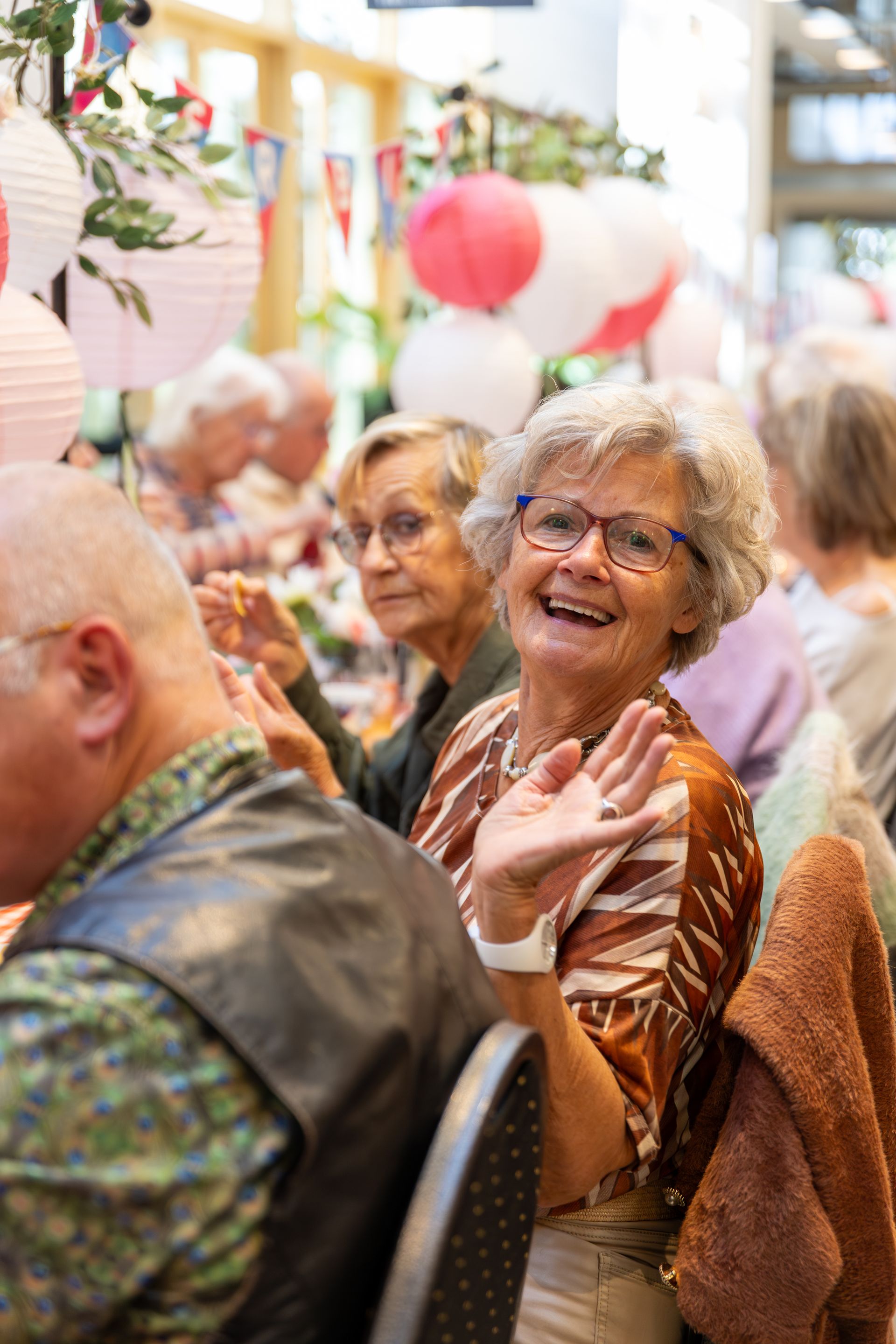 Een vrouw zwaait naar een tafel vol mensen. De kamer is versierd met roze en witte ballonnen.