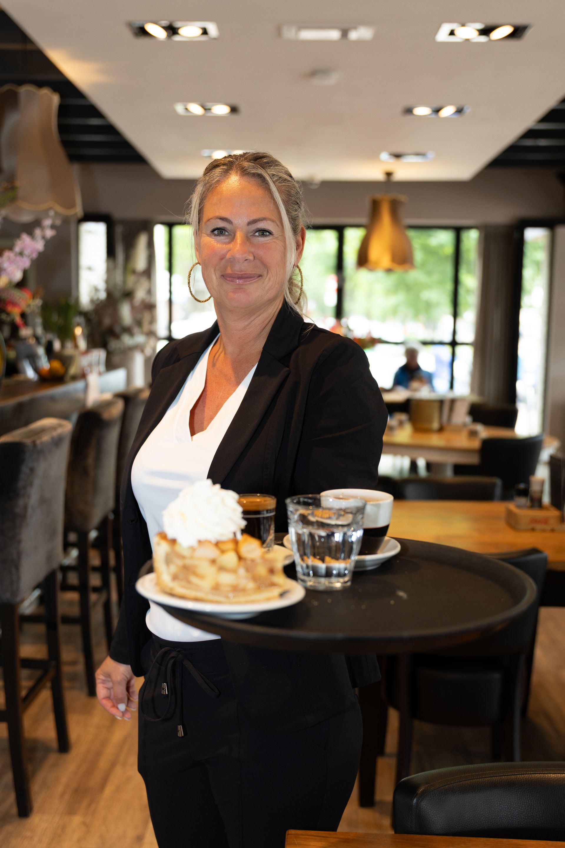 Vrouw in een restaurant met een dienblad met appeltaart en koffie; ze lacht naar de camera.