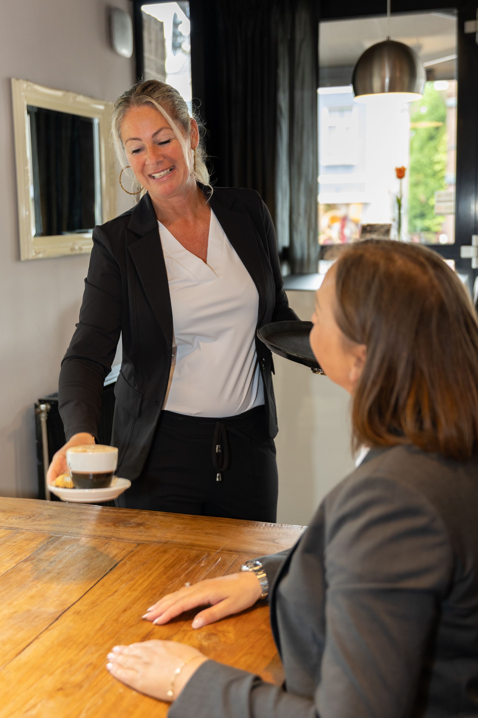 Een vrouw in een pak serveert koffie aan een andere vrouw aan een houten tafel.