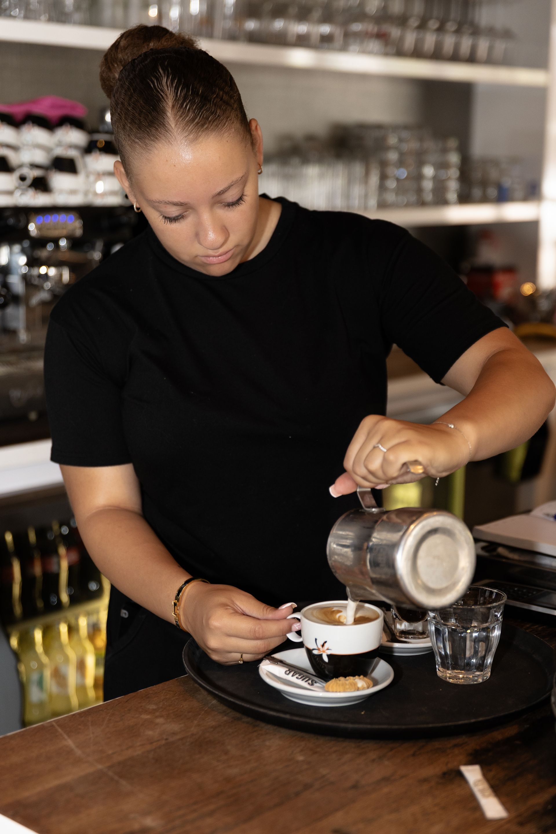 Een vrouw schenkt koffie uit een metalen pot in een kopje op een dienblad, in een caféomgeving.