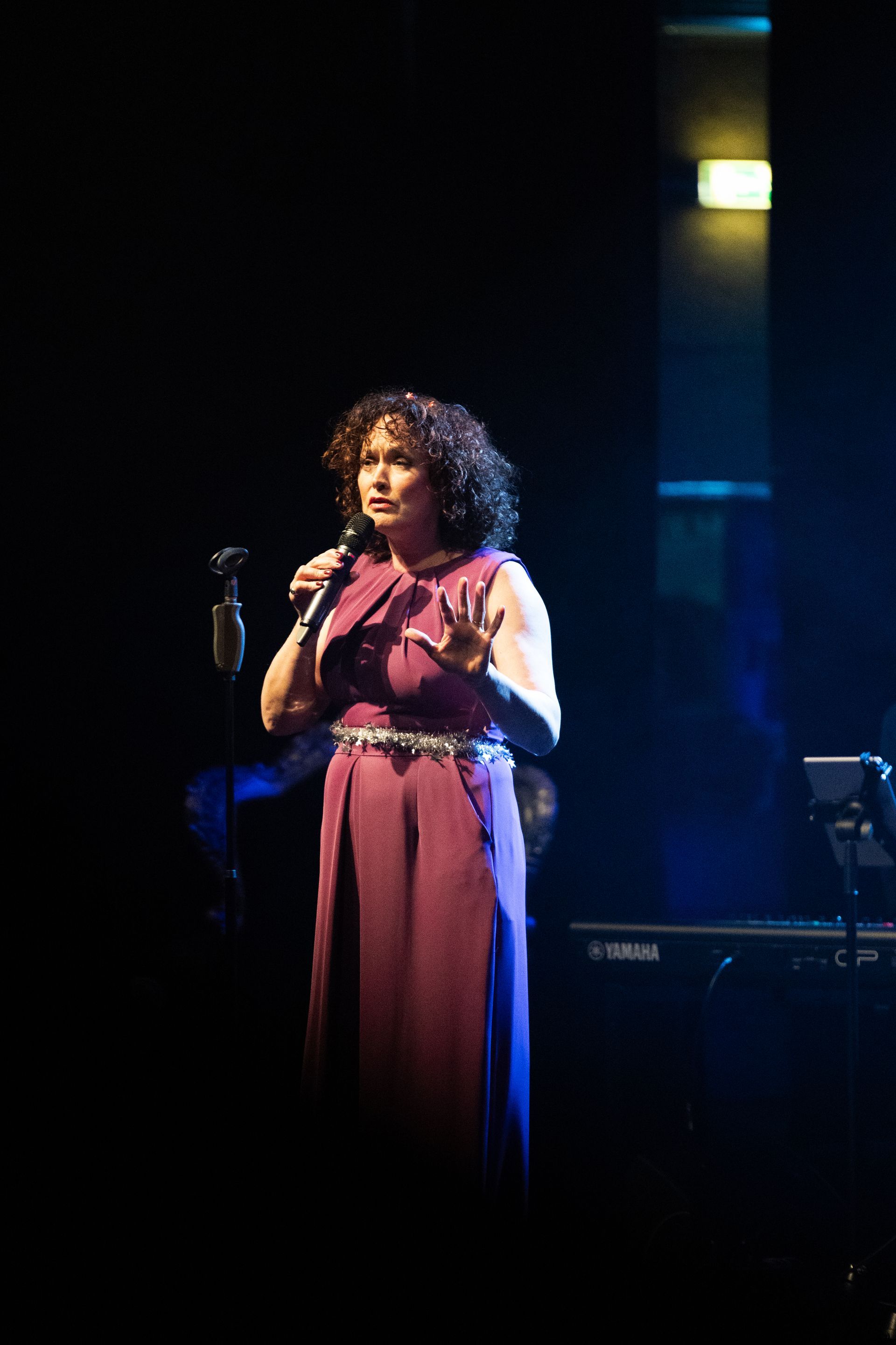 Een vrouw zingt op het podium, gekleed in een pruimkleurige jurk, met een microfoon in haar hand.