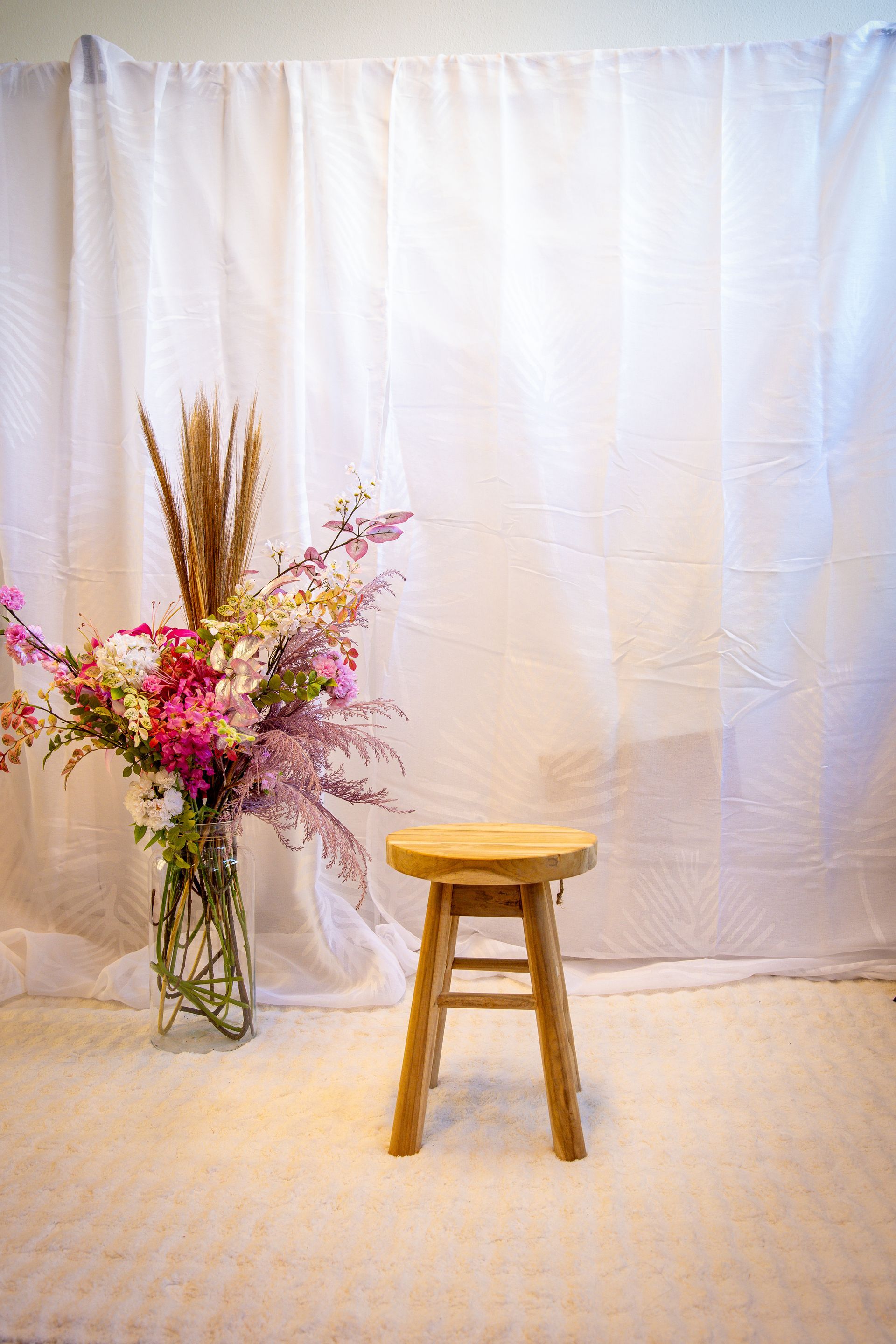 Een klein houten krukje naast een glazen vaas met roze bloemen, tegen een achtergrond van een wit gordijn.