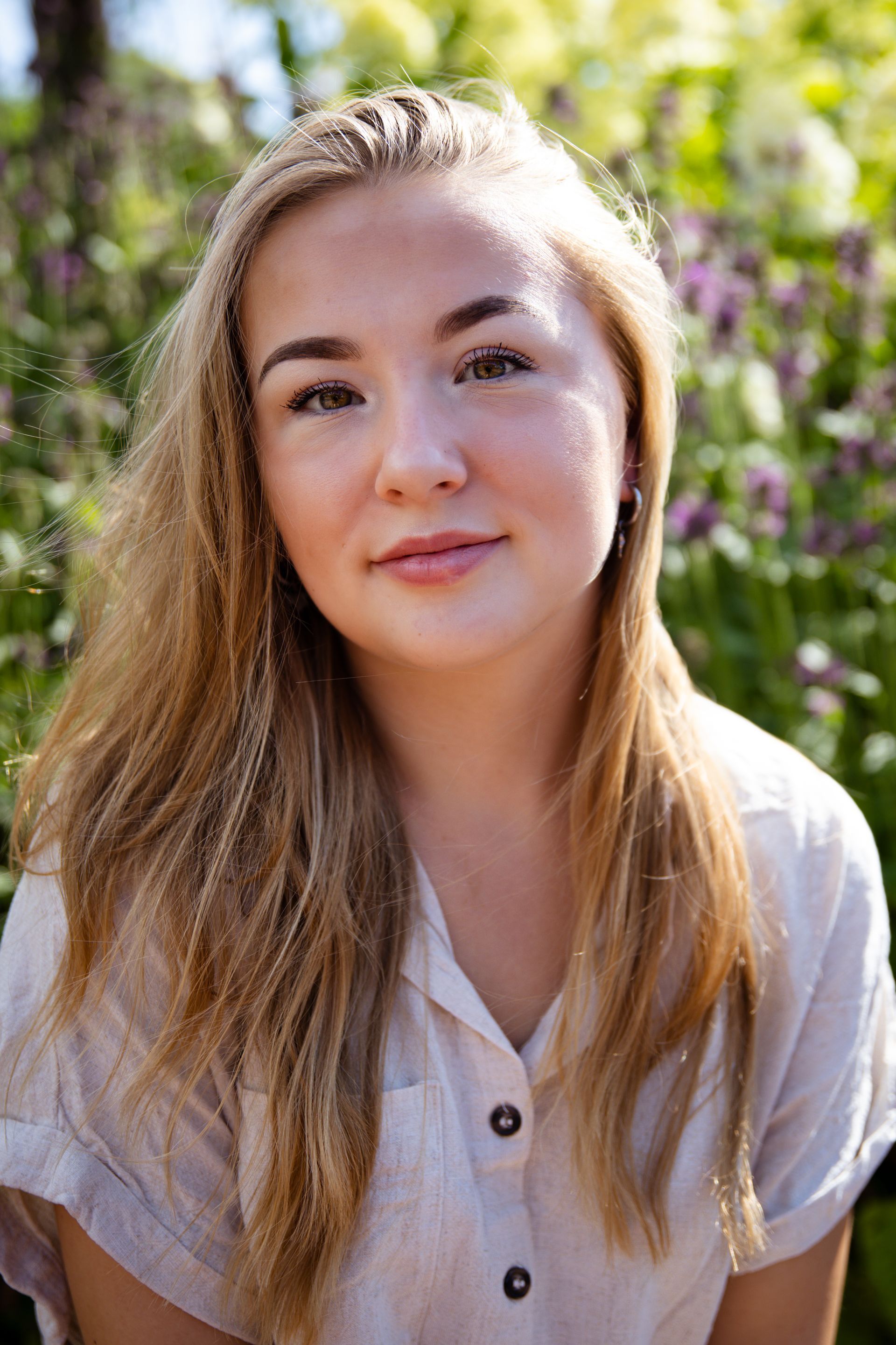 Jonge vrouw met blond haar in een lichtgekleurd topje, licht glimlachend. Buiten, omringd door groen en bloemen.