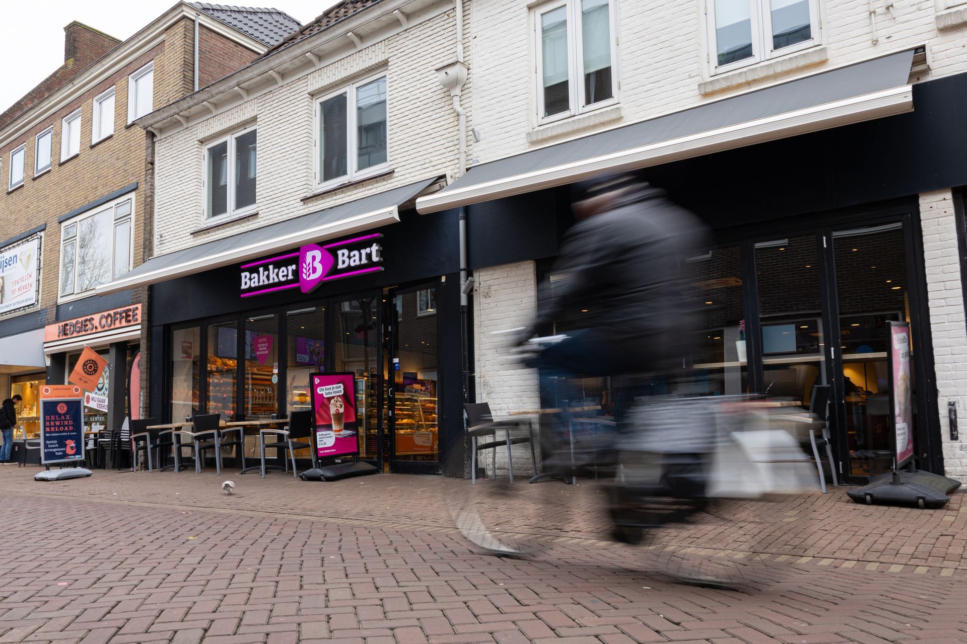 Bakkerijgevel met een wazige fietser die voorbijrijdt op een bakstenen straat.