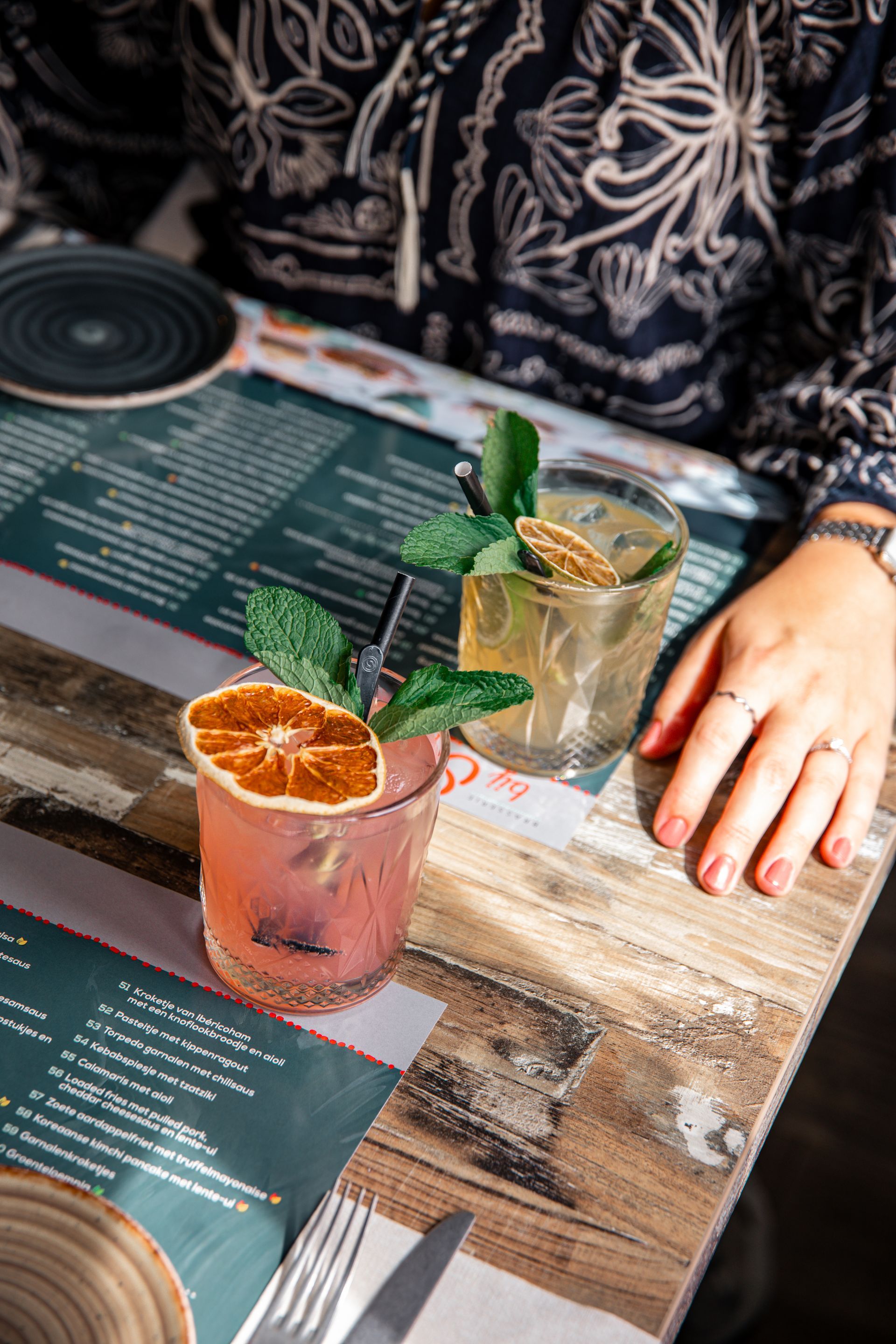 Twee cocktails, gegarneerd met citrus en munt, staan ​​op een houten tafel. De handen van een persoon zijn zichtbaar.