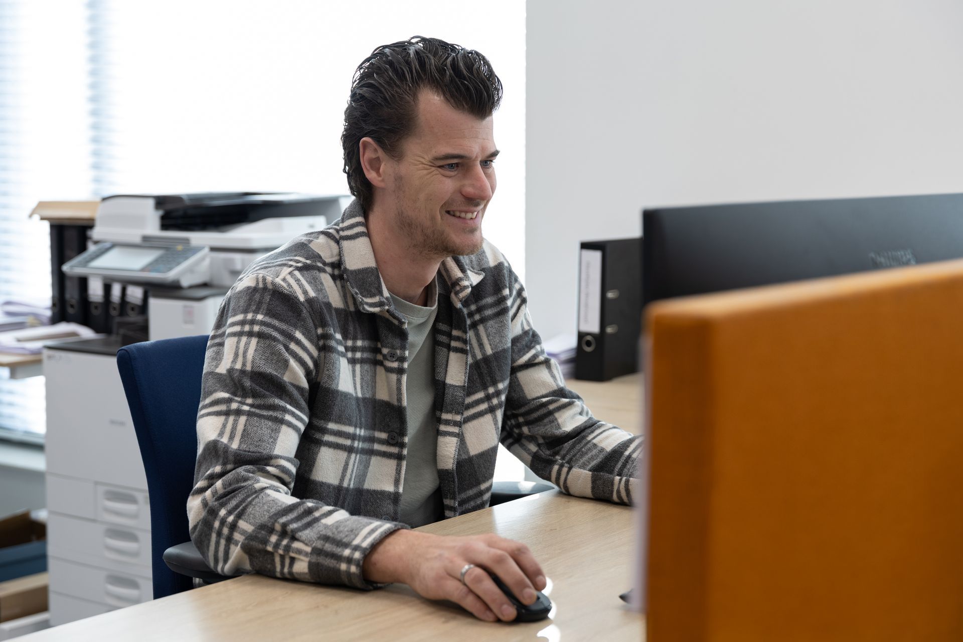 Een man glimlacht terwijl hij achter een computer zit aan een bureau in een kantooromgeving. 