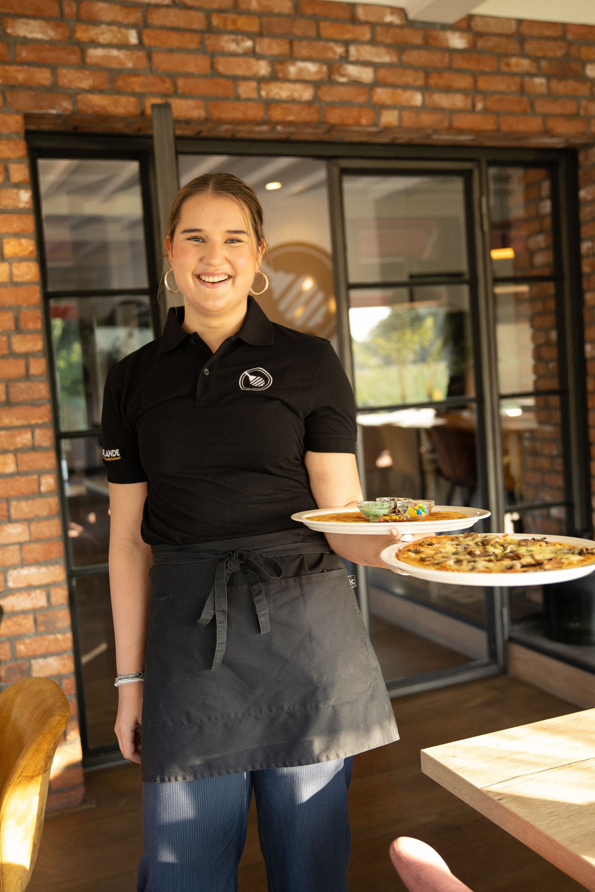 Een ober in een zwart uniform draagt ​​twee pizza's en staat glimlachend bij de ingang van een restaurant.