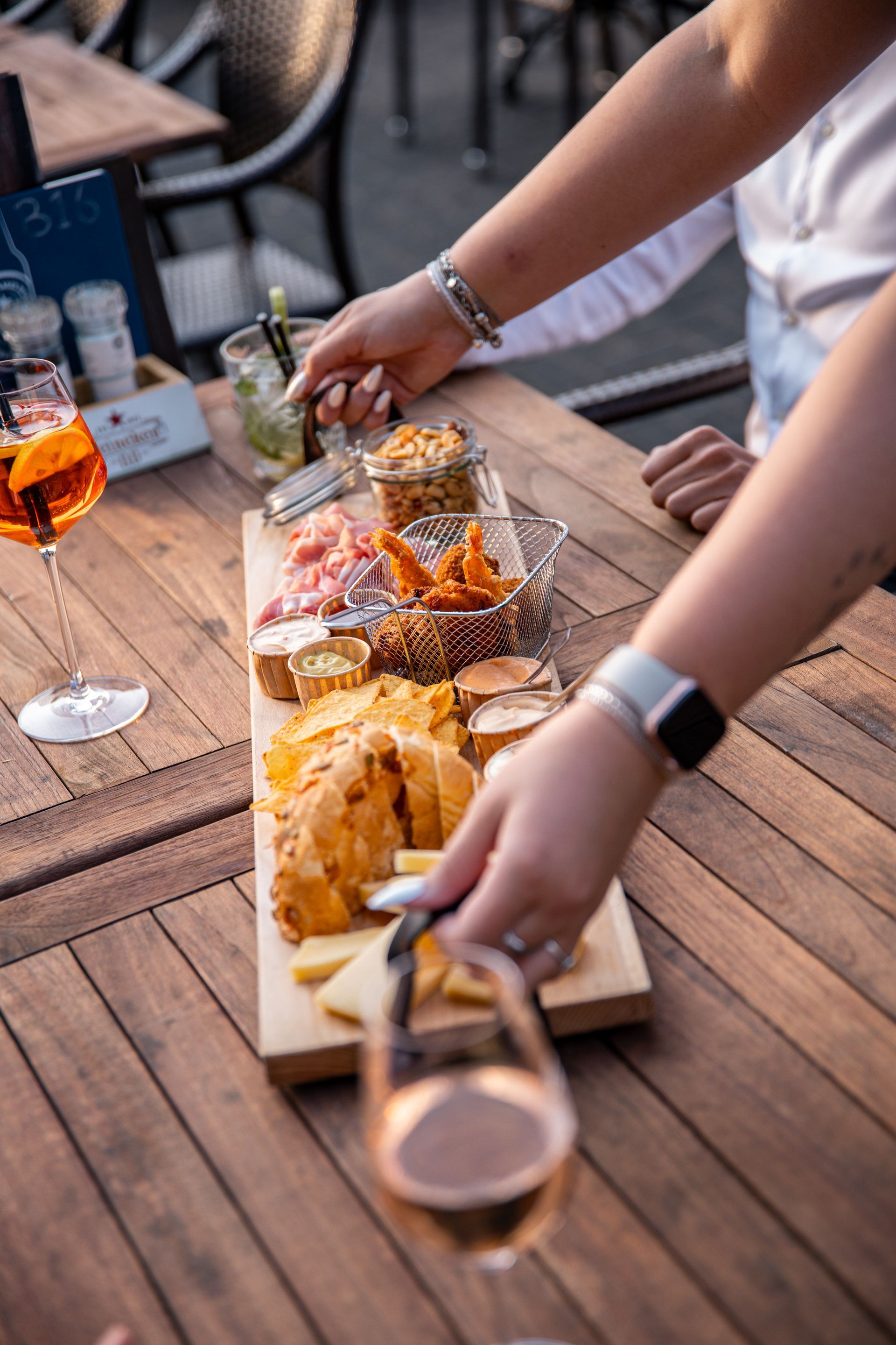 Iemand die een charcuterieplank op een houten tafel buiten klaarmaakt, met een Aperol Spritz erbij.