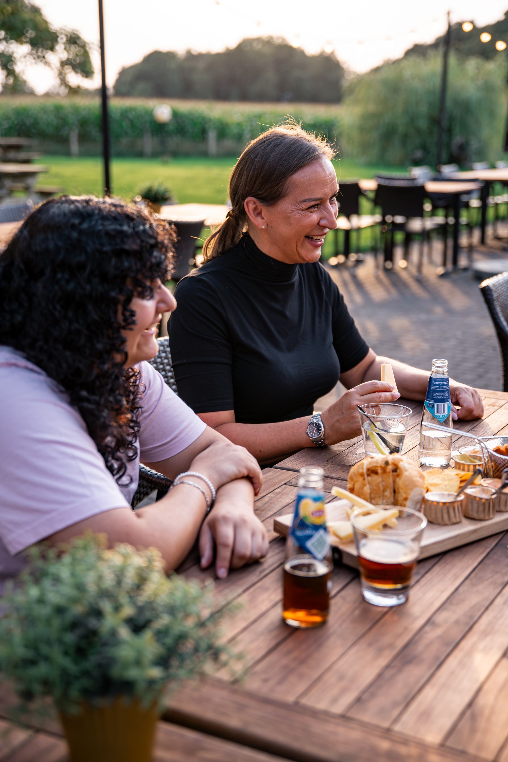Twee vrouwen zitten met een glimlach aan een tafeltje buiten, met eten en drinken.