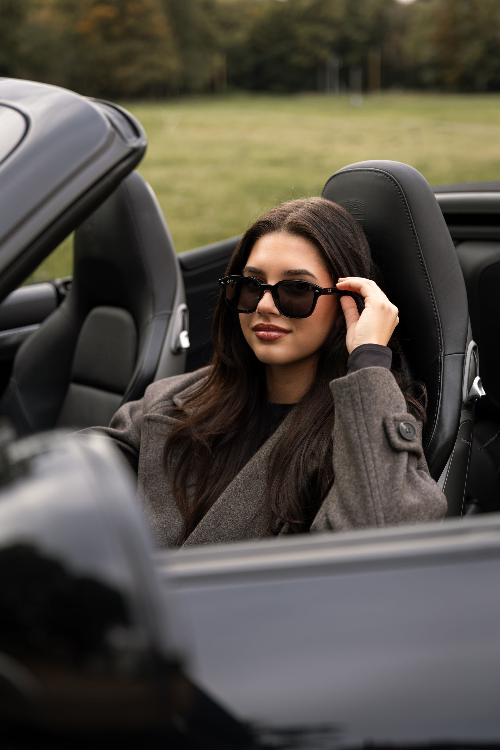 Vrouw in een grijze jas met zonnebril zit in een zwarte cabriolet.