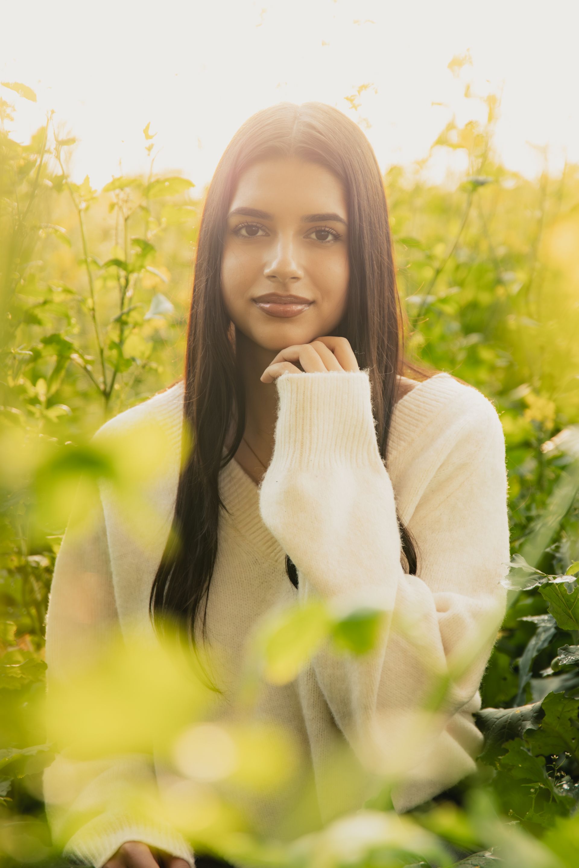 Een vrouw in een witte trui poseert in een veld met groene planten, waar het zonlicht doorheen filtert.