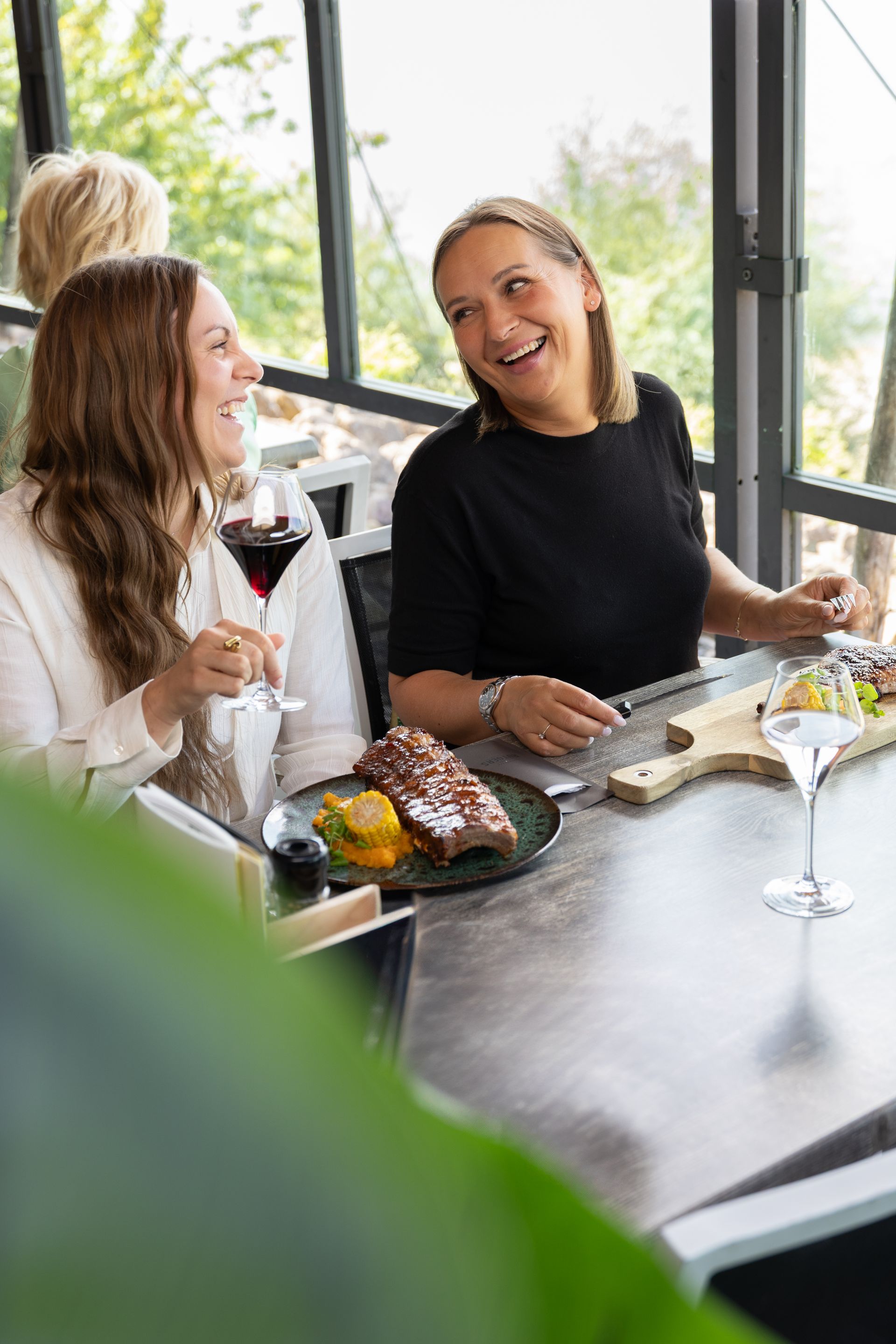 Twee vrouwen zitten lachend aan een tafel, terwijl ze eten en wijn drinken in een modern, licht restaurant.