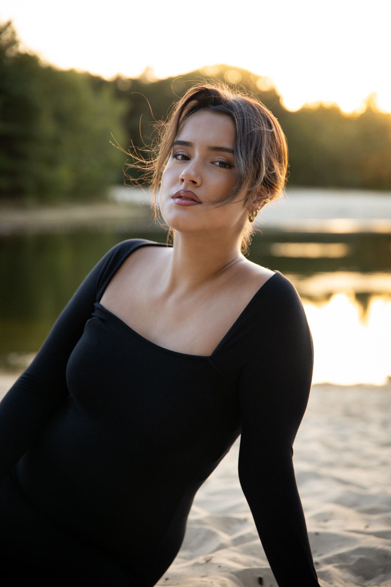 Vrouw in zwarte jurk, zittend op het zand bij het water, kijkt naar de camera; zonsondergang op de achtergrond.
