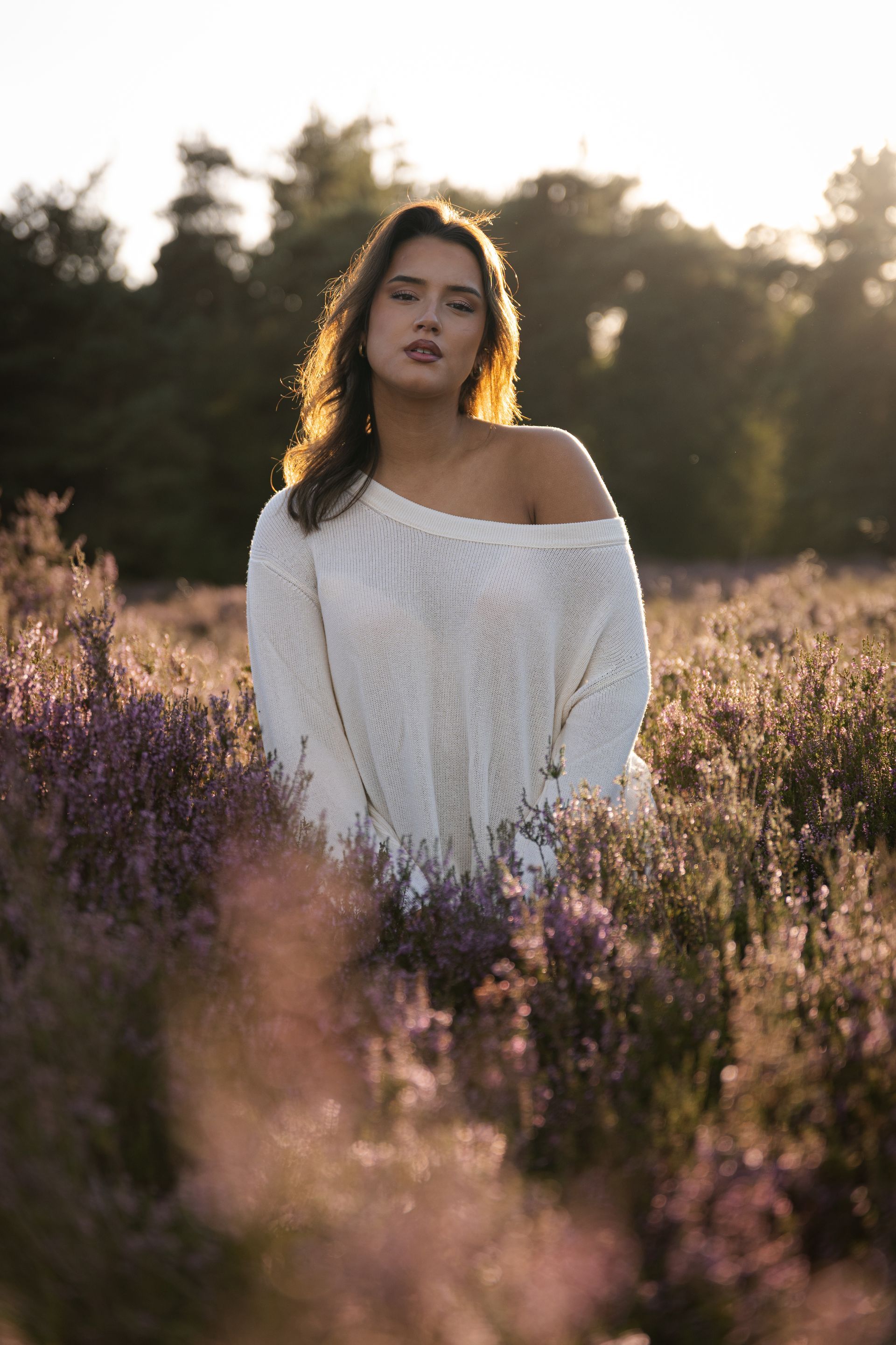 Een vrouw in een witte trui staat in een veld met paarse bloemen, haar rug wordt verlicht door de zon.
