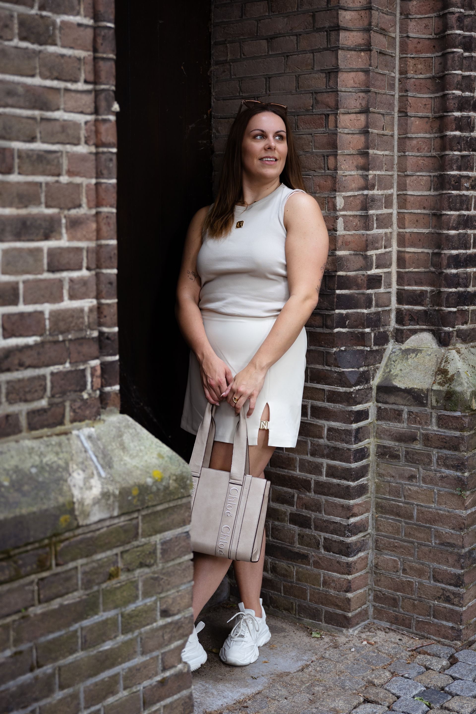 Een vrouw in een crèmekleurige outfit, met een tas in haar hand, staat in een bakstenen deuropening en kijkt niet in beeld.
