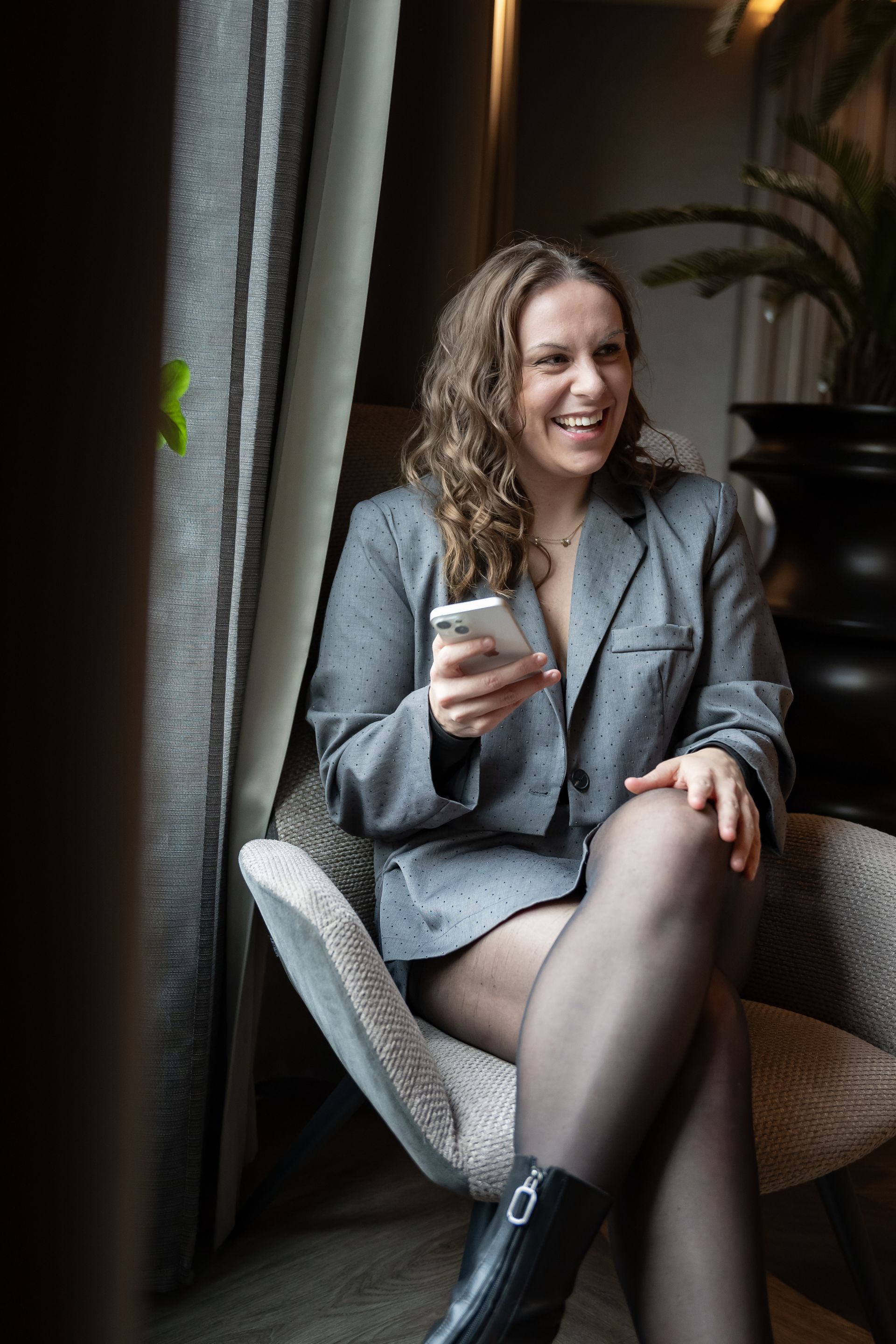 Vrouw in grijze blazer, met telefoon in de hand, glimlacht terwijl ze in een stoel bij een raam zit.