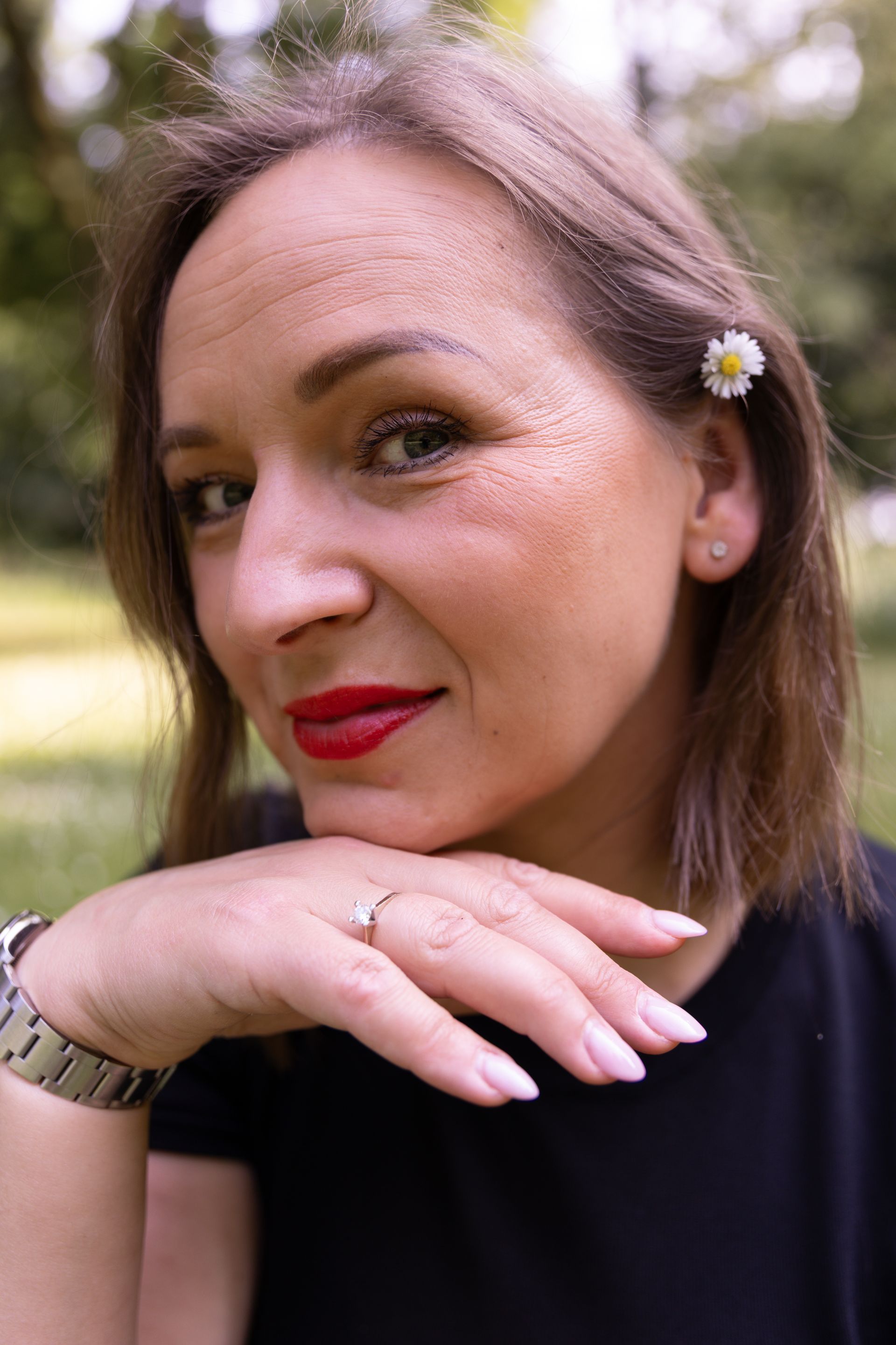 Vrouw met rode lippenstift, hand onder de kin, klein madeliefje in het haar, lachend in de buitenlucht.