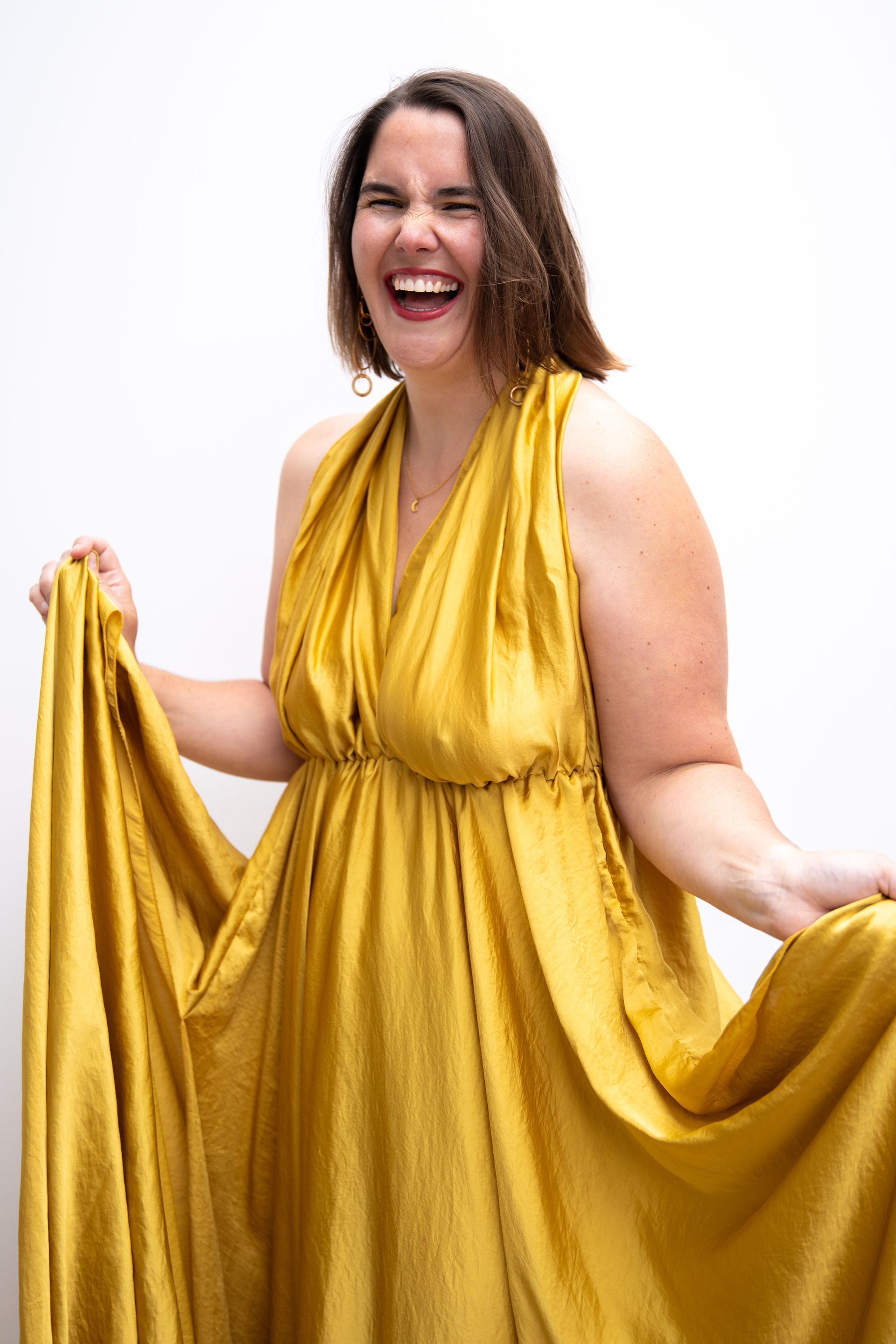 Een lachende vrouw, gekleed in een zwierige gouden jurk, houdt de stof omhoog tegen een witte achtergrond.
