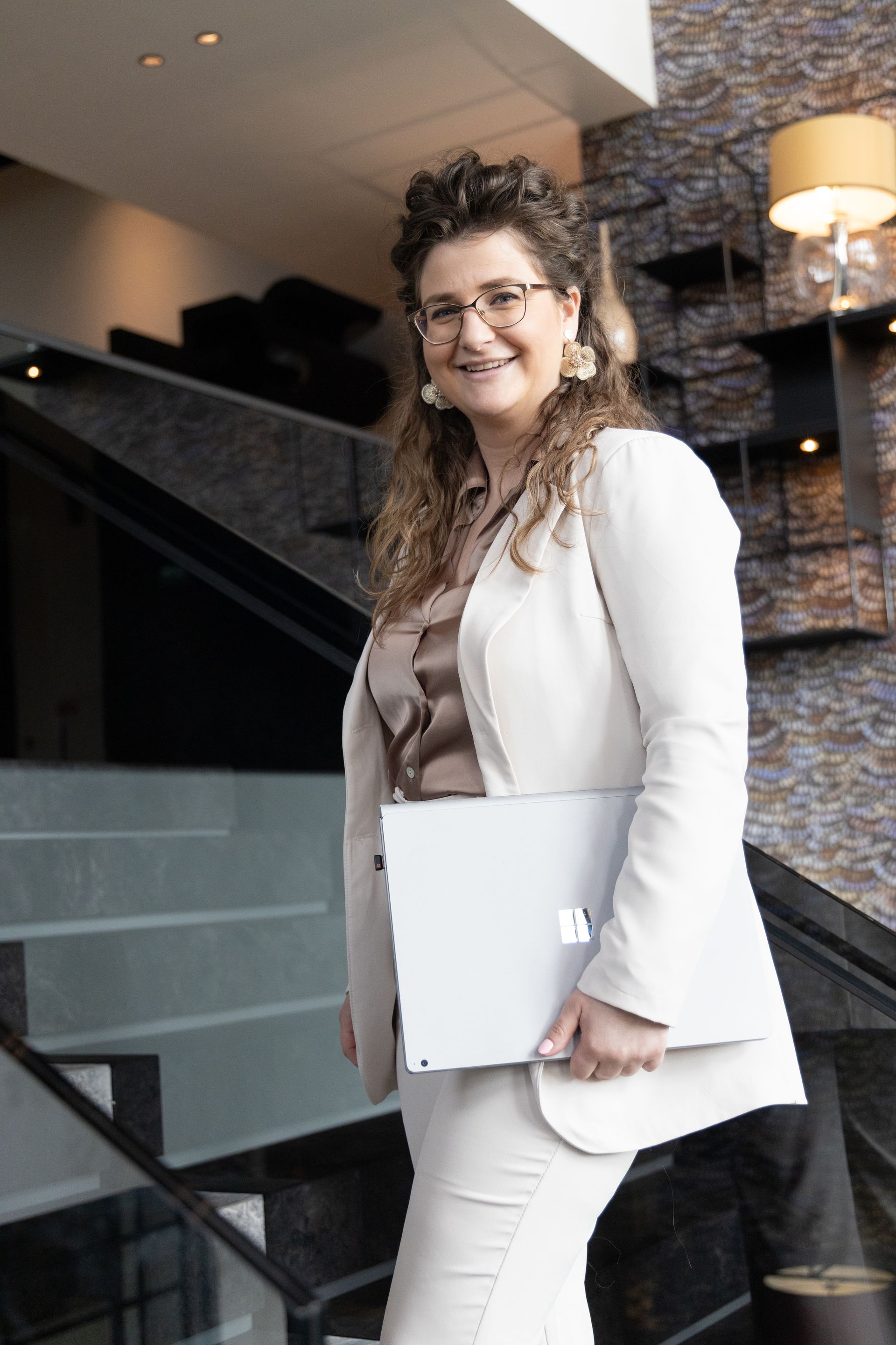 Een vrouw in een crèmekleurig broekpak staat op de trap met een laptop in haar handen. 