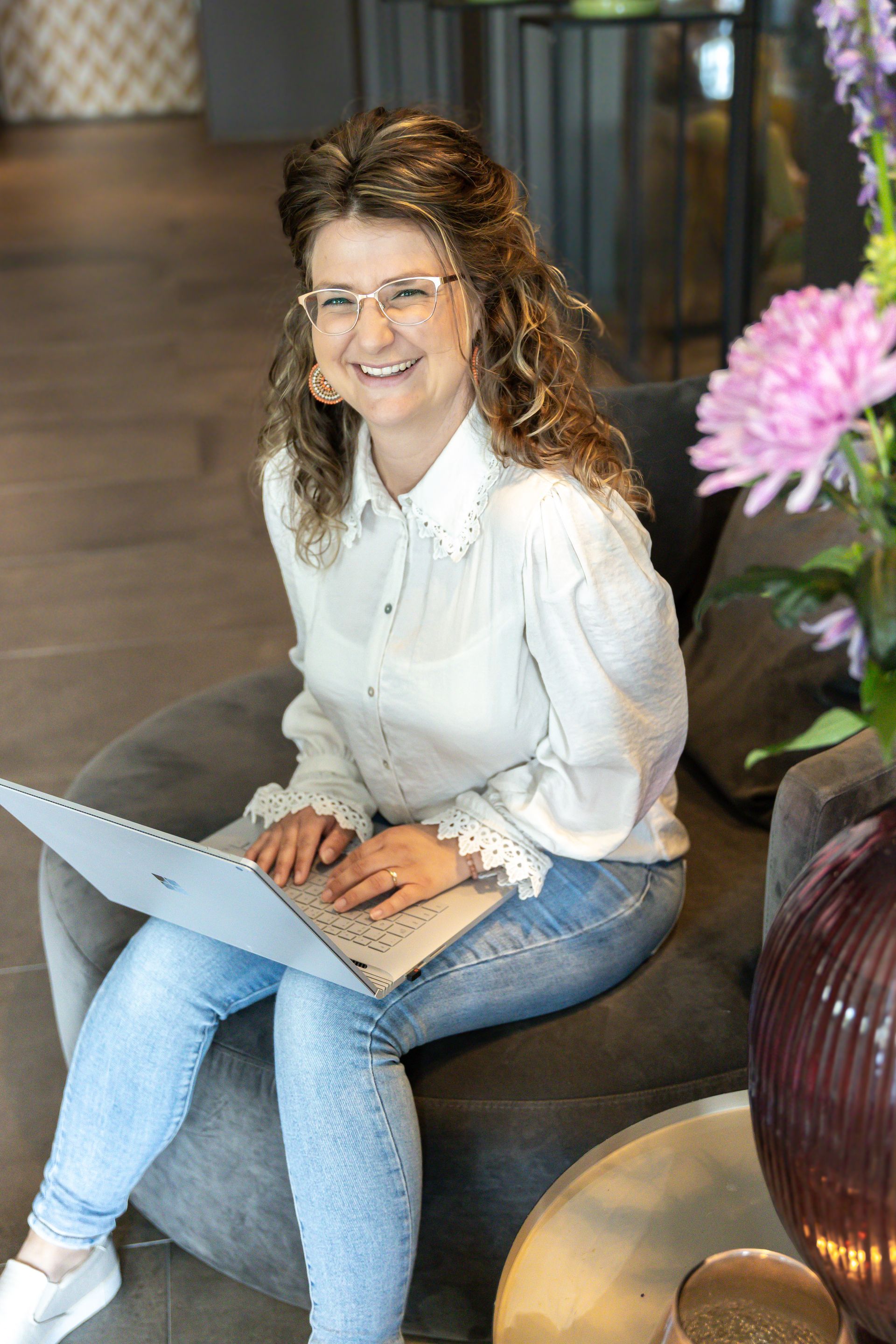 Vrouw in witte blouse en jeans glimlacht terwijl ze op een laptop zit in een fauteuil.