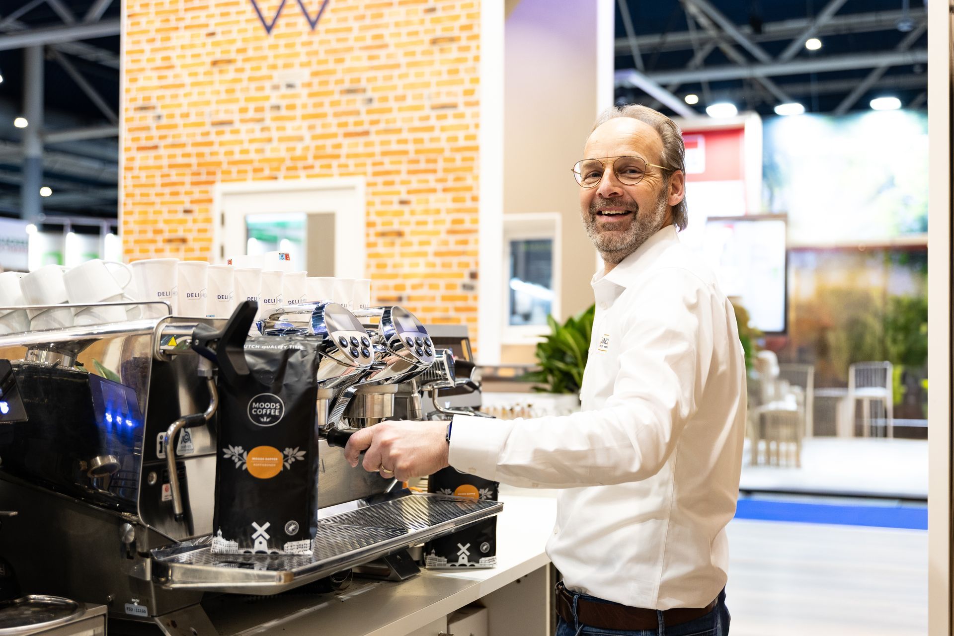 Een lachende man bedient een espressomachine op een beursstand met een bakstenen muur als achtergrond.