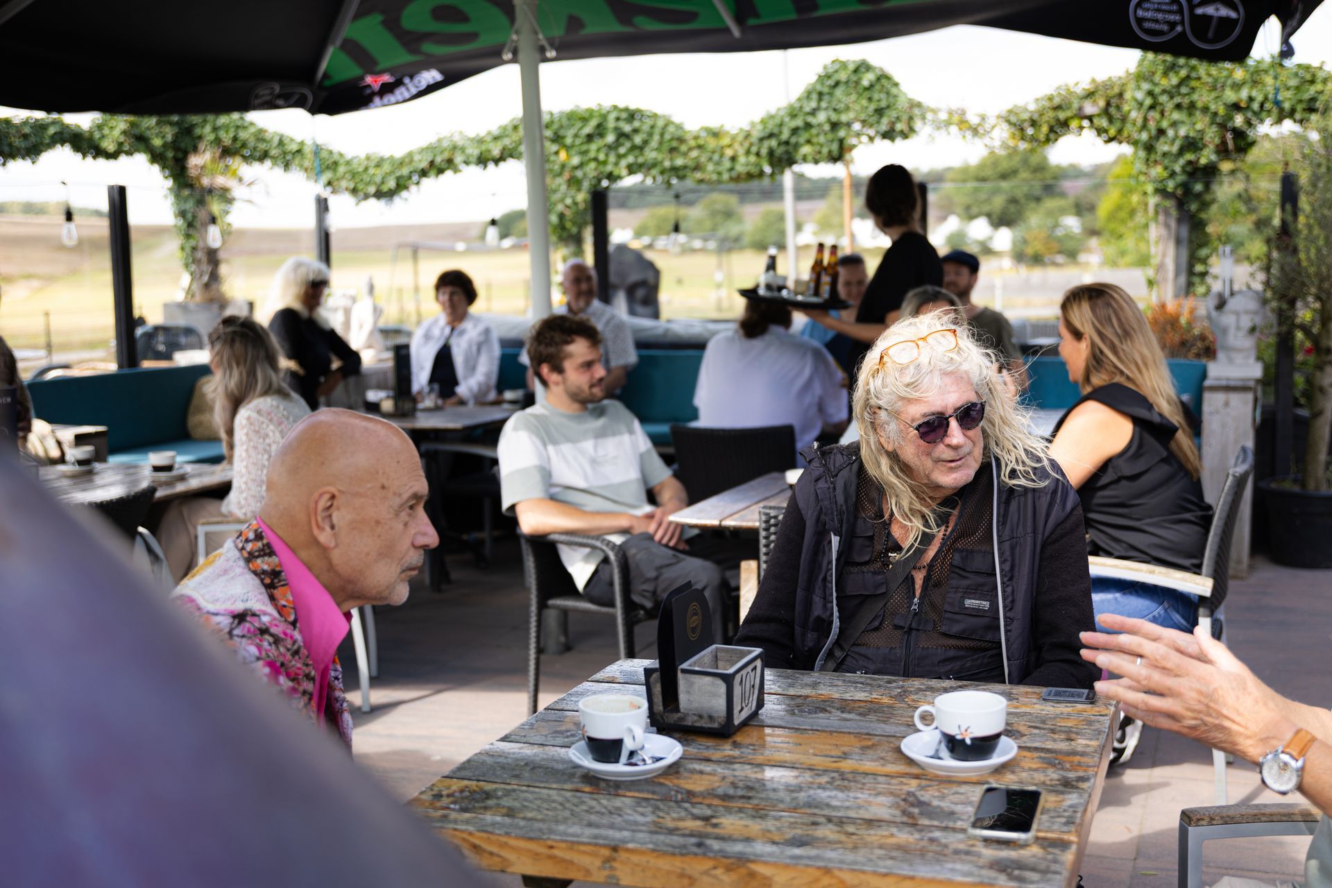 Mensen zitten aan tafels op een terras. Eén persoon draagt ​​een zonnebril. Anderen drinken koffie en voeren een gesprek.